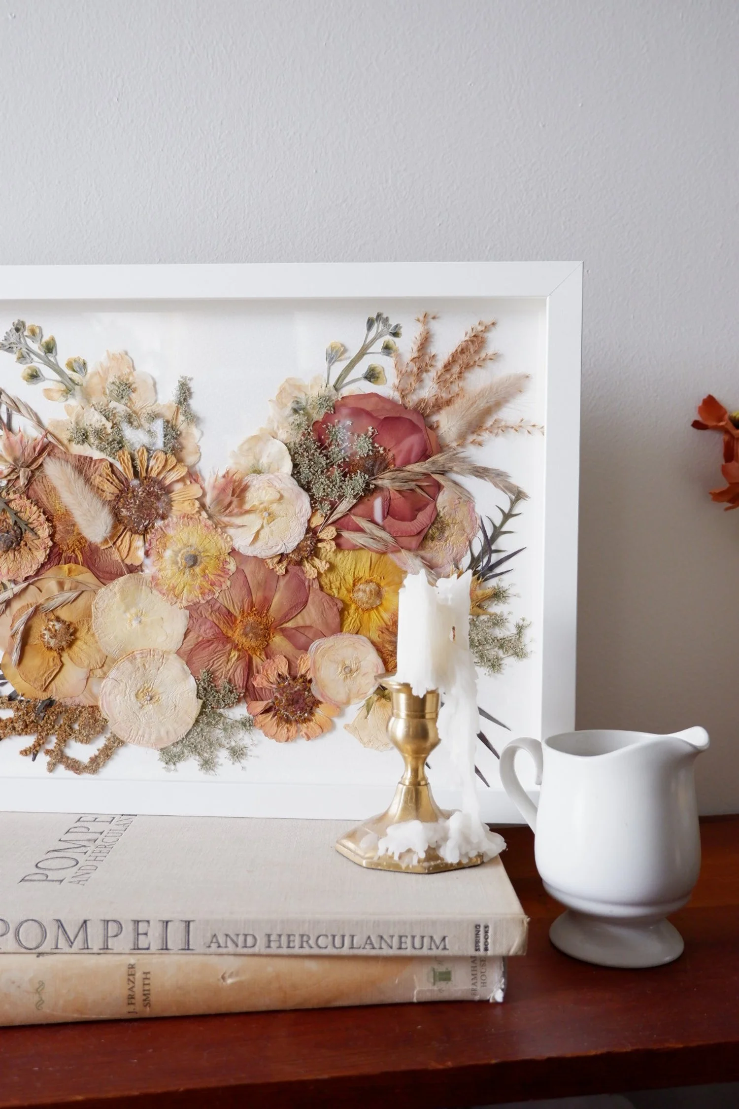 Dried floral arrangement in a white shadow box frame, a lit white candle in a gold candle holder with wax dripping, a white ceramic pitcher, and two books on a wooden surface.