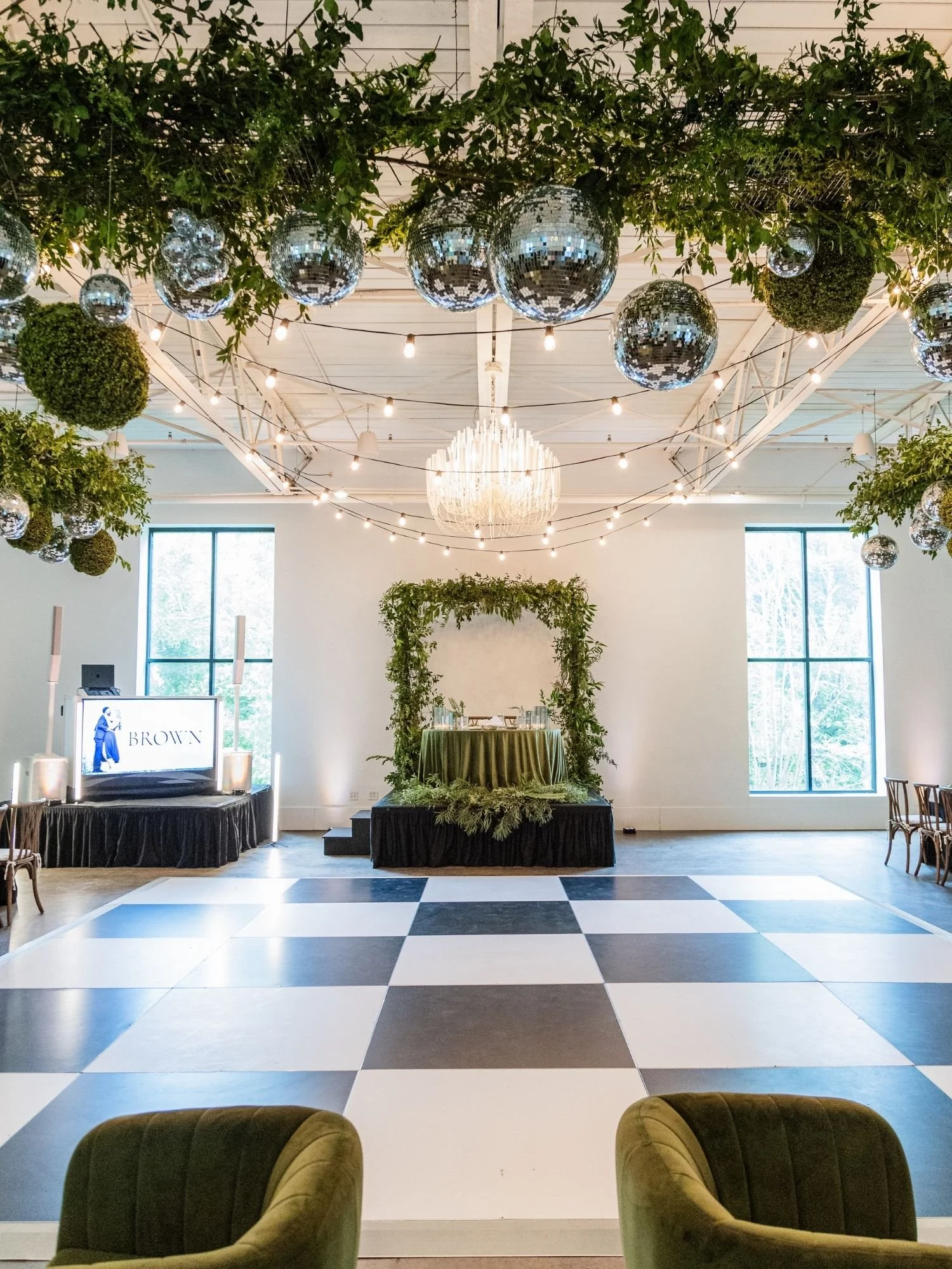 checkerboard dance floor with sweetheart table in behind it- decorated with a large greenery arch and greenery at base of table. Large greenery and disco ball chandelier above and dj in the corner of dancefloor.