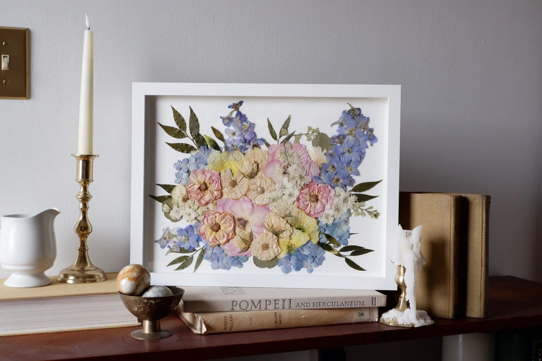 Decorative framed pressed flower arrangement with pink, yellow, white, and blue flowers and green leaves, on a wooden shelf with books, a candlestick, a jug, a bowl with stones, and candles.