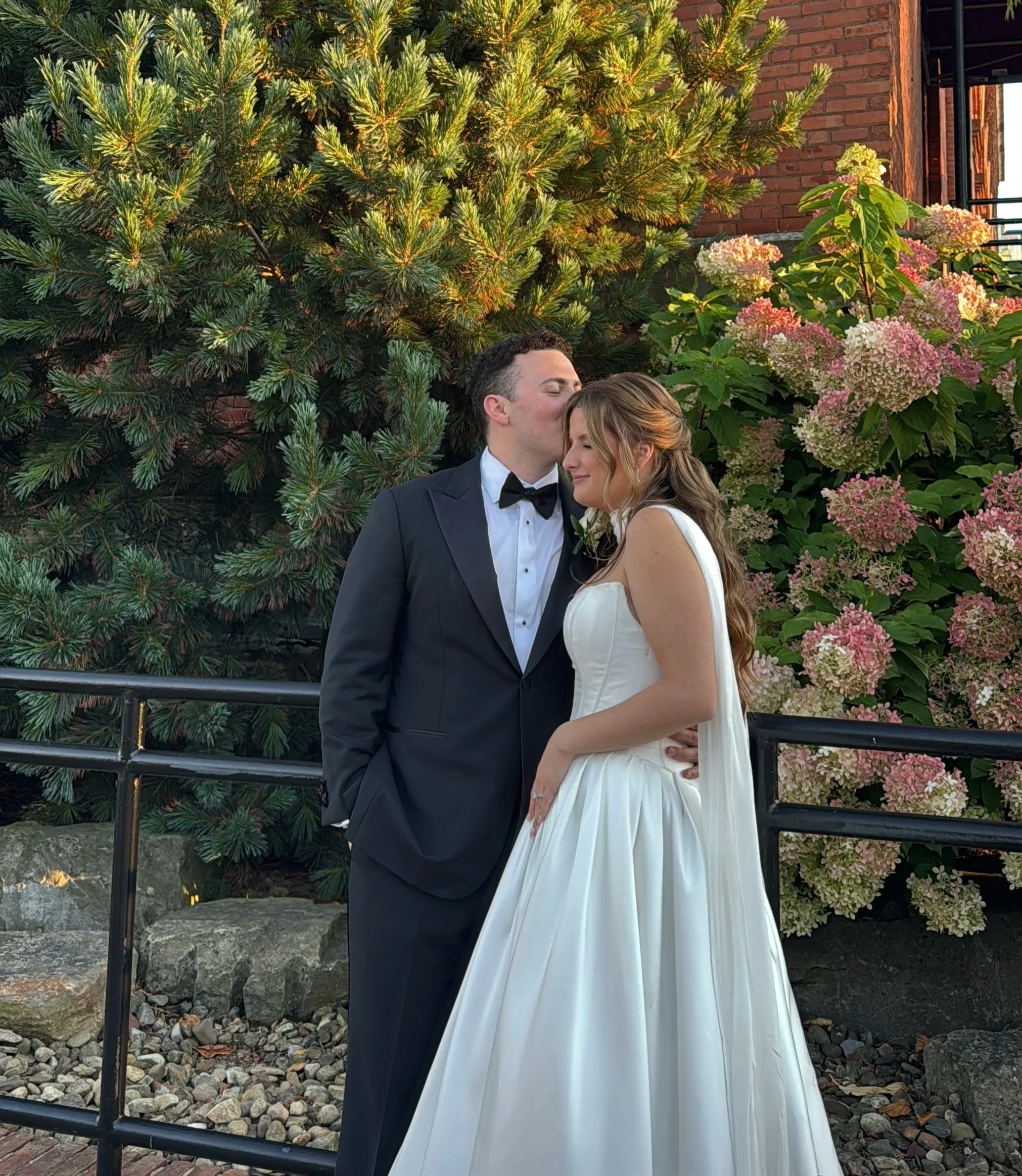 A groom and bride sharing a kiss in front of lush greenery and pink hydrangea flowers, outside a brick building.