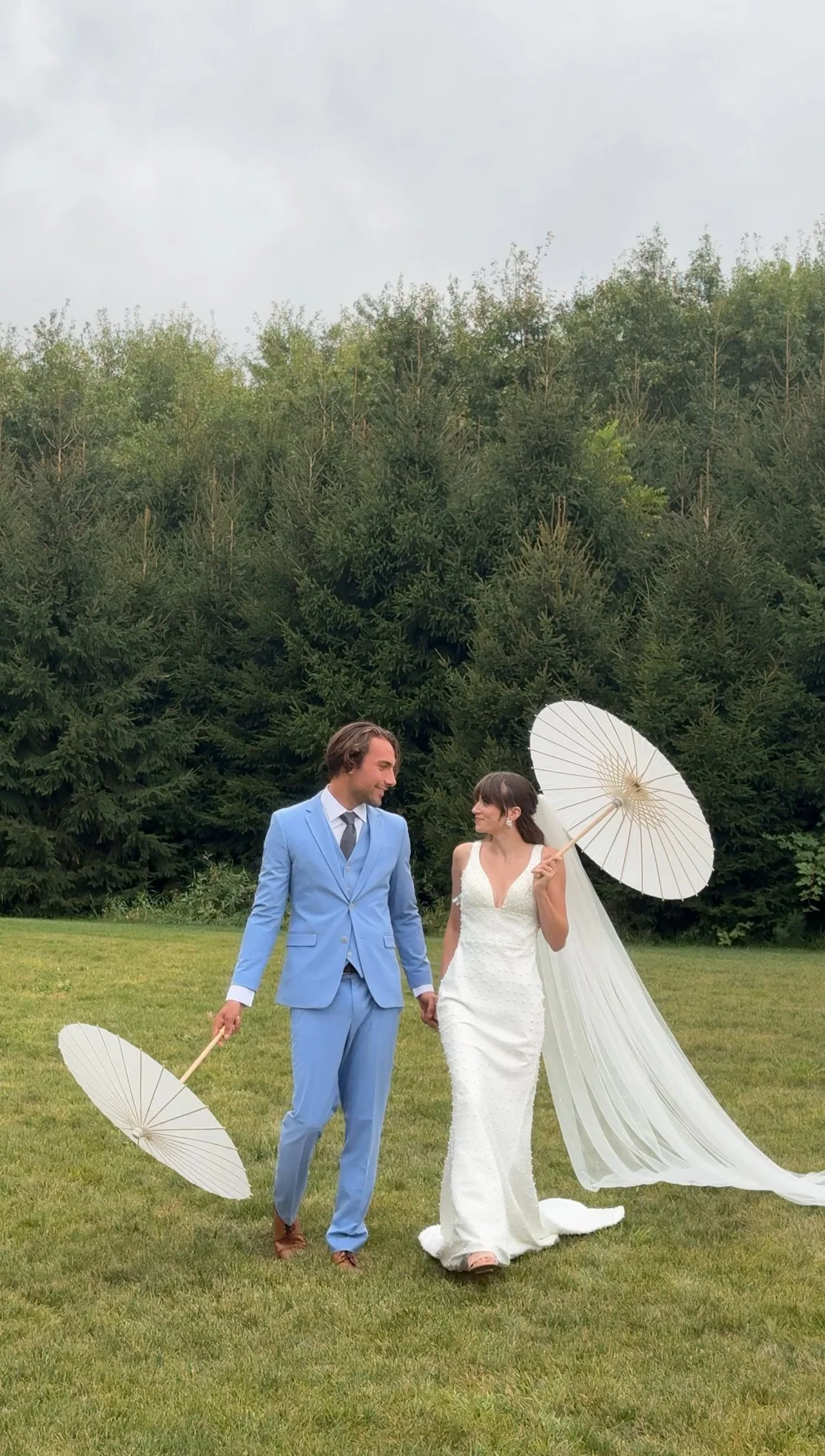 Bride and groom walking hand-in-hand outdoors, both holding white parasols, bride wearing a white gown with long train and veil, groom in a light blue suit, greenery and trees in the background.