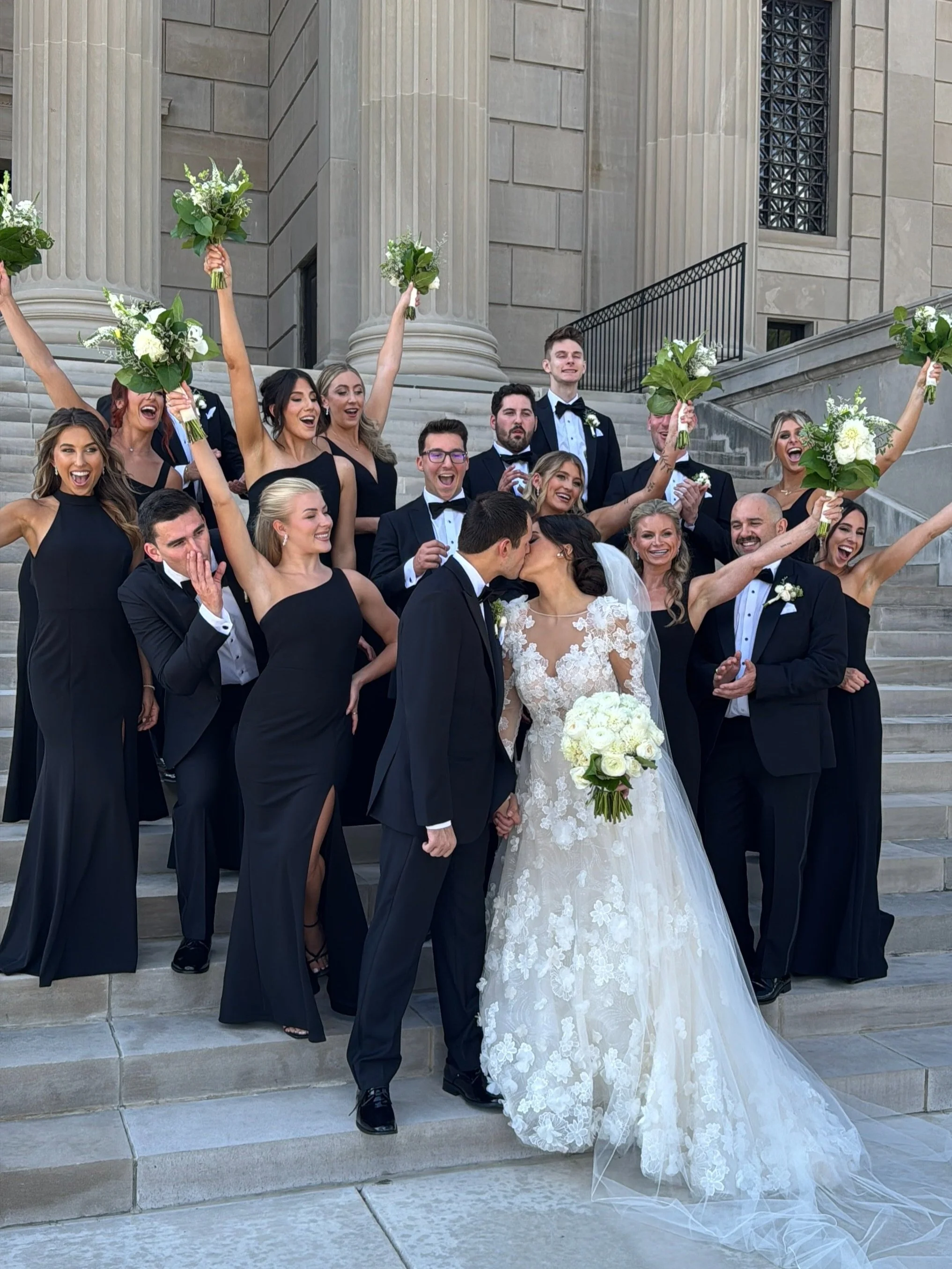 A wedding party on the steps of a courthouse or church, with the bride and groom kissing in the center, surrounded by bridesmaids and groomsmen celebrating with bouquets and smiles.