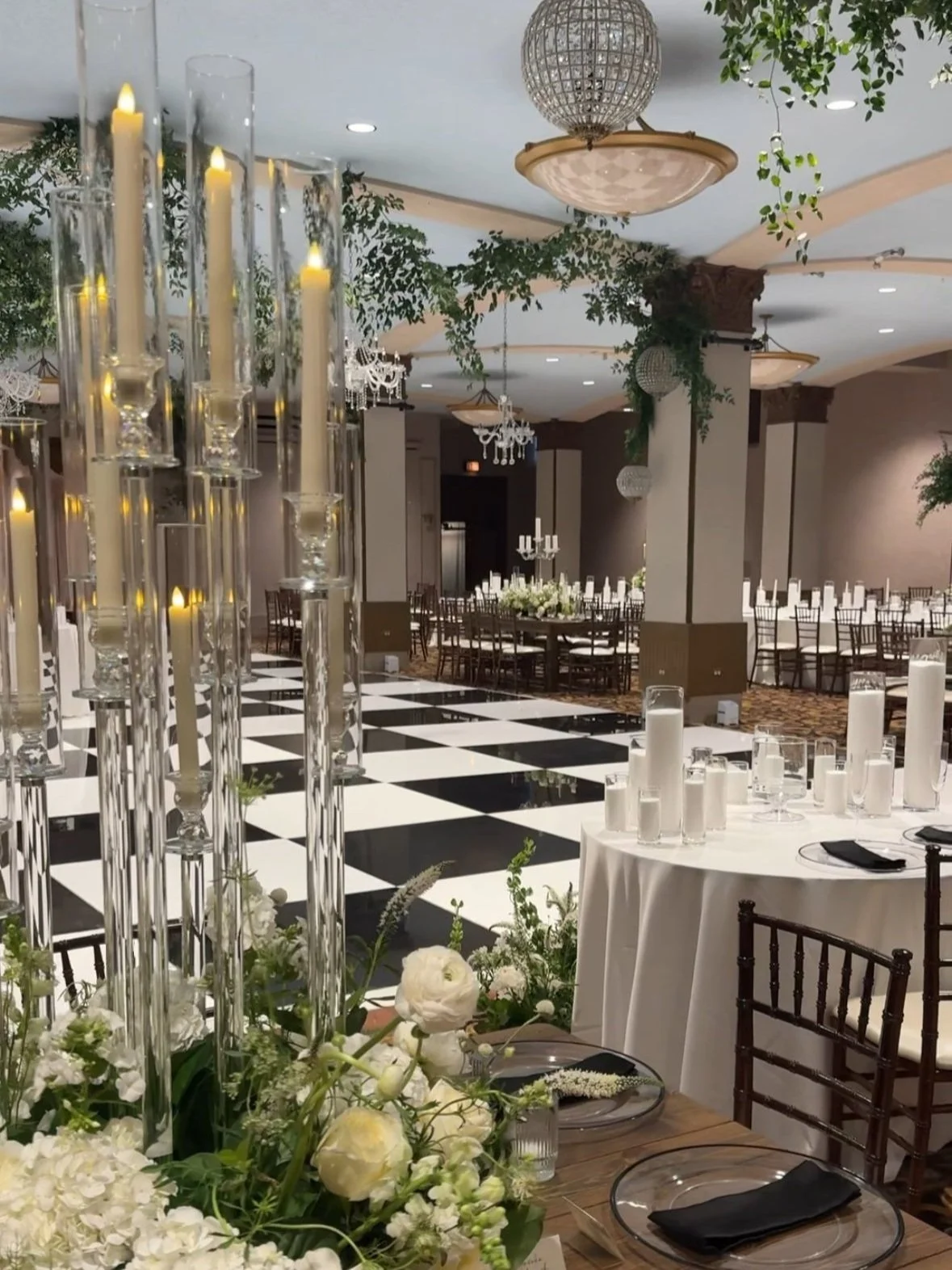 Elegant banquet hall decorated for a wedding or special event with black and white checkered dance floor, tall candle centerpieces, floral arrangements, and multiple dining tables set with black napkins.