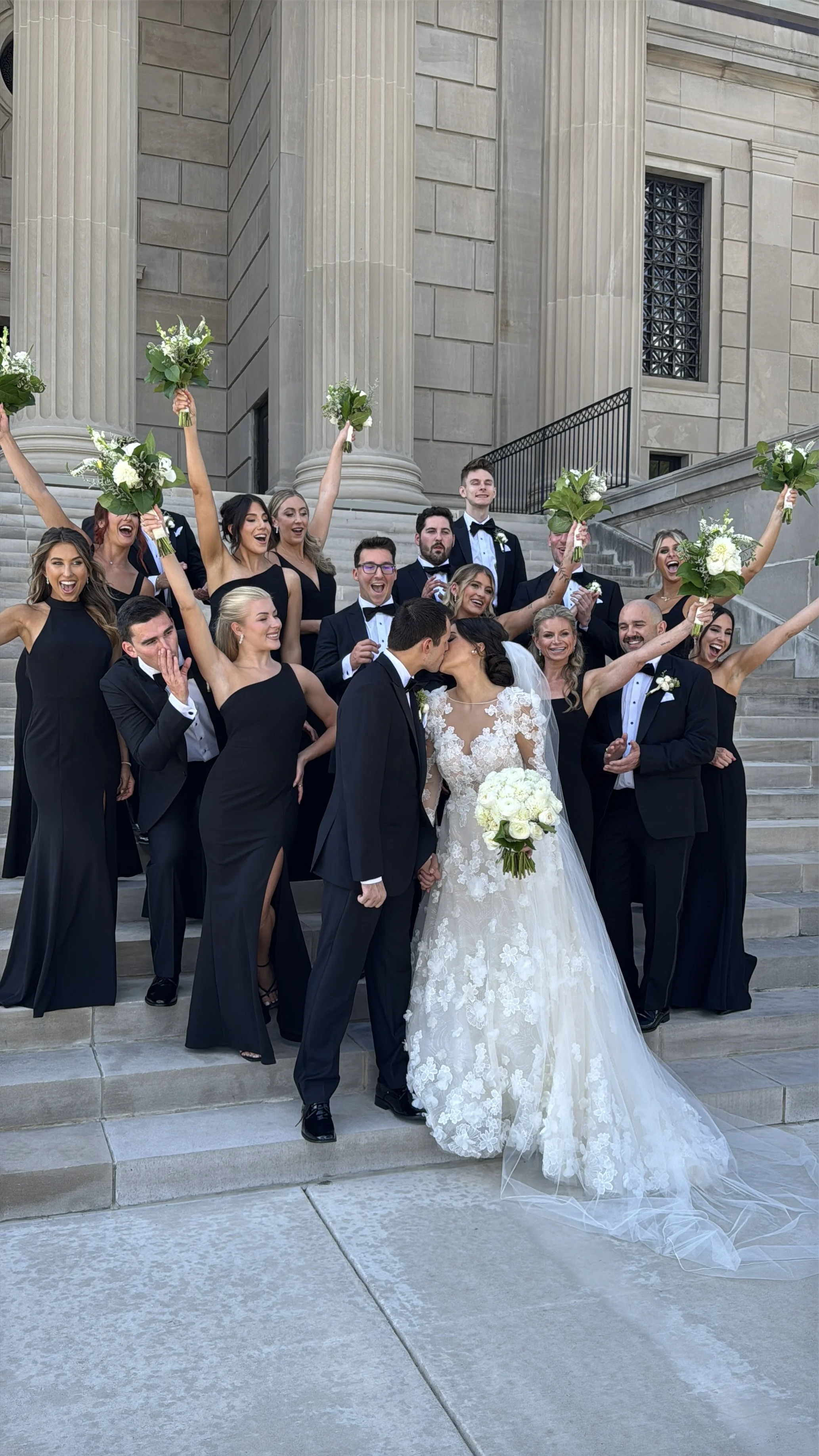 A group of wedding guests, dressed in formal black attire, celebrating on the steps outside a large, neoclassical building with tall columns. A bride and groom are kissing at the front, with the bride holding a bouquet of white flowers, while the guests cheer and raise their own bouquets.