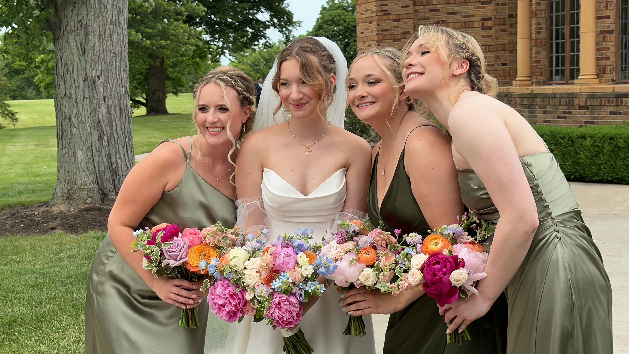 Four women in formal dresses pose for a photo outdoors, with a large bouquet of colorful flowers in front of them. One woman is wearing a white bridal gown, suggesting a wedding celebration, while the other three are in matching emerald green dresses, possibly bridesmaids, standing on green grass near a brick building and trees.