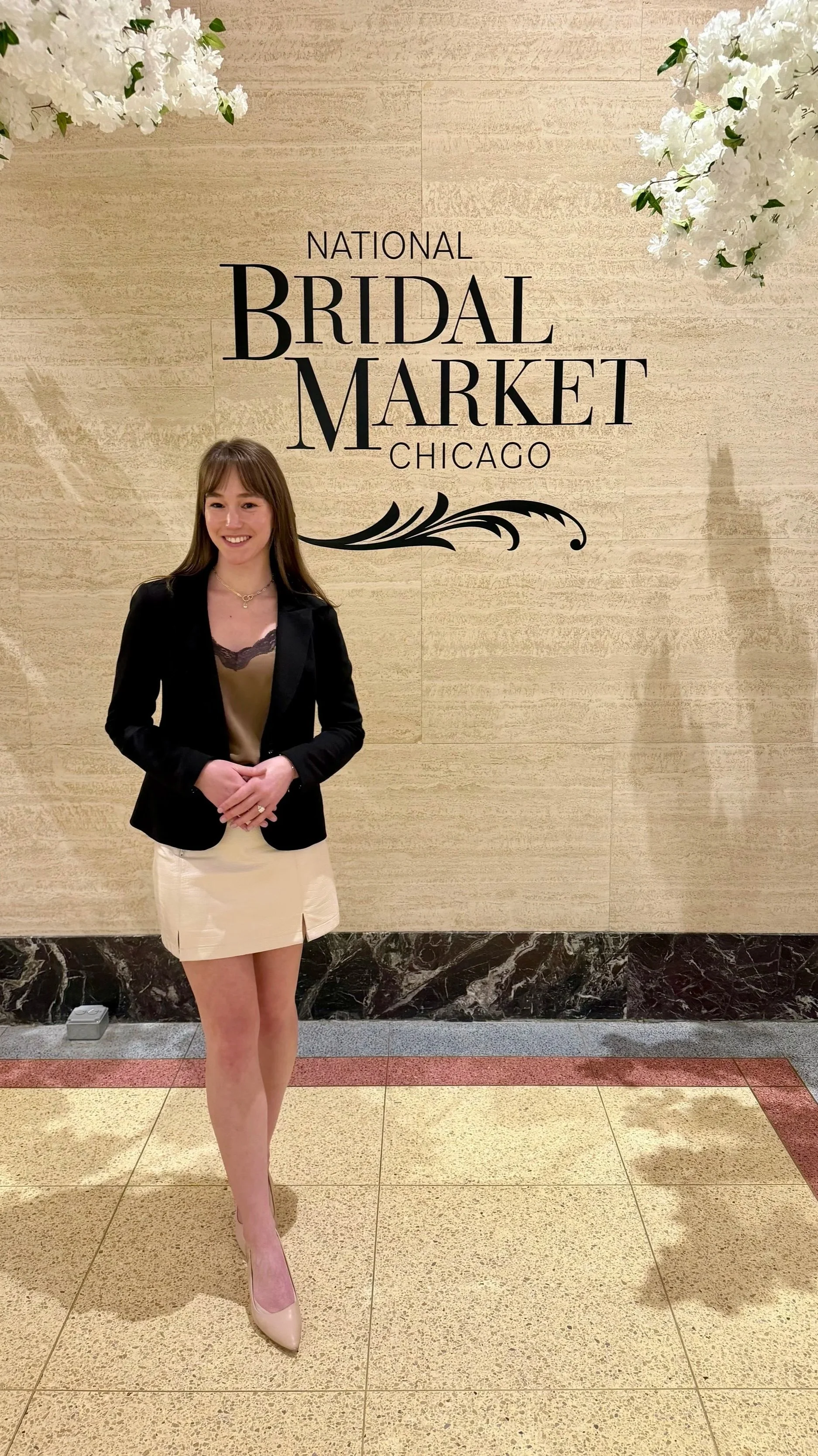 A woman in a black blazer and beige skirt posing in front of a sign that reads 'National Bridal Market Chicago' with white floral decorations overhead.