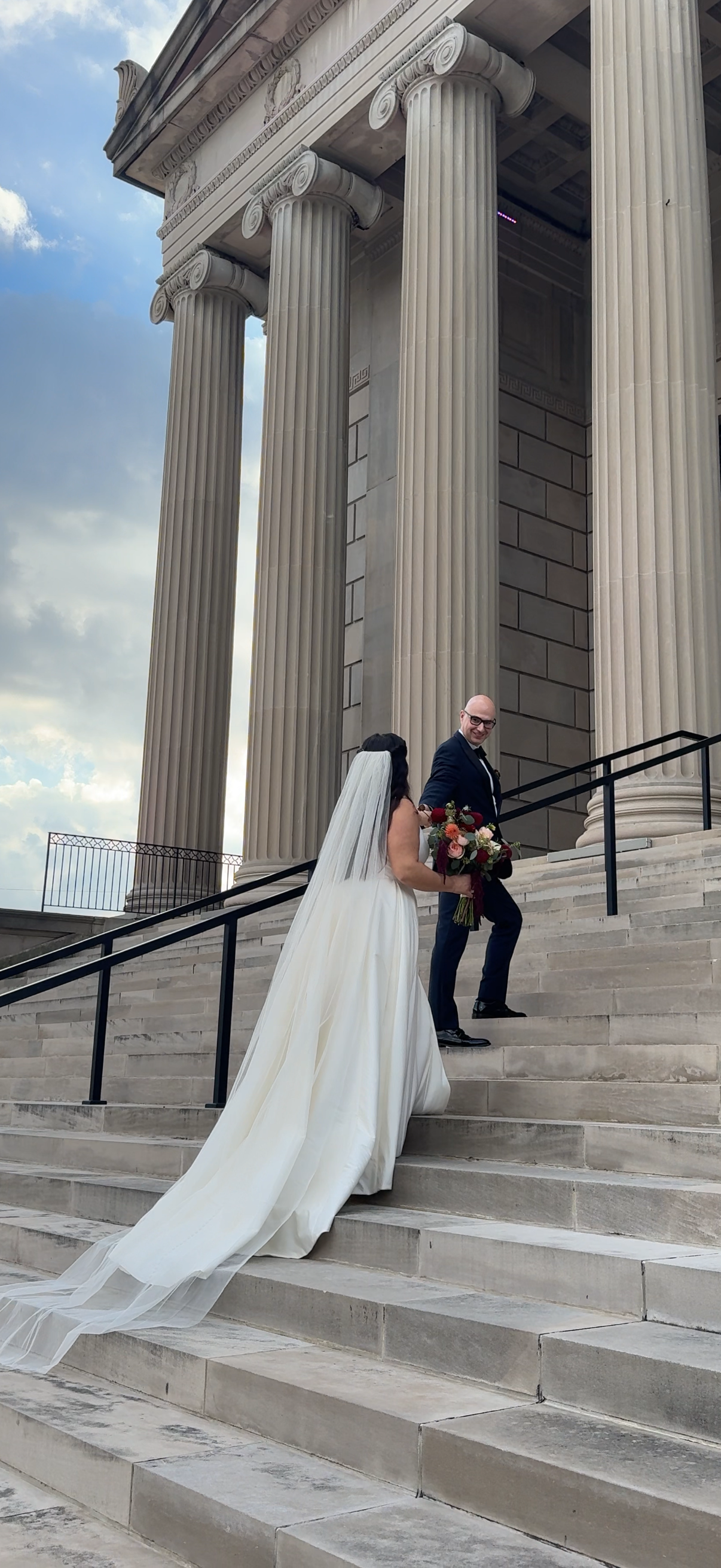 A bride and groom descending the steps of a classical building with large columns, the bride in a long white wedding dress holding a bouquet, and the groom in a dark suit smiling