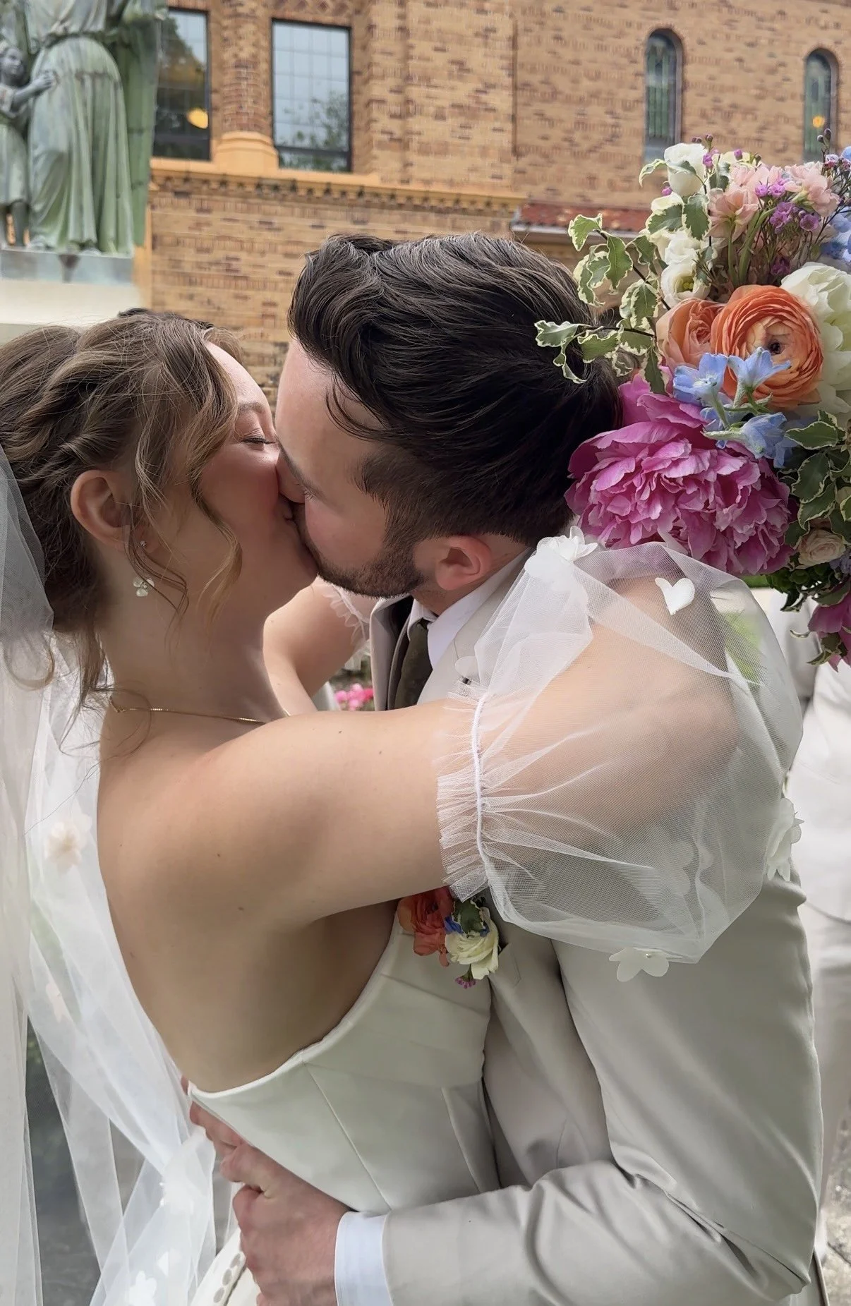 A bride and groom sharing a kiss outdoors, with the bride in a white dress and veil, holding a bouquet of pink, purple, white, and peach flowers. The groom is wearing a light-colored suit and bow tie. Behind them is a brick building and a statue of a woman.