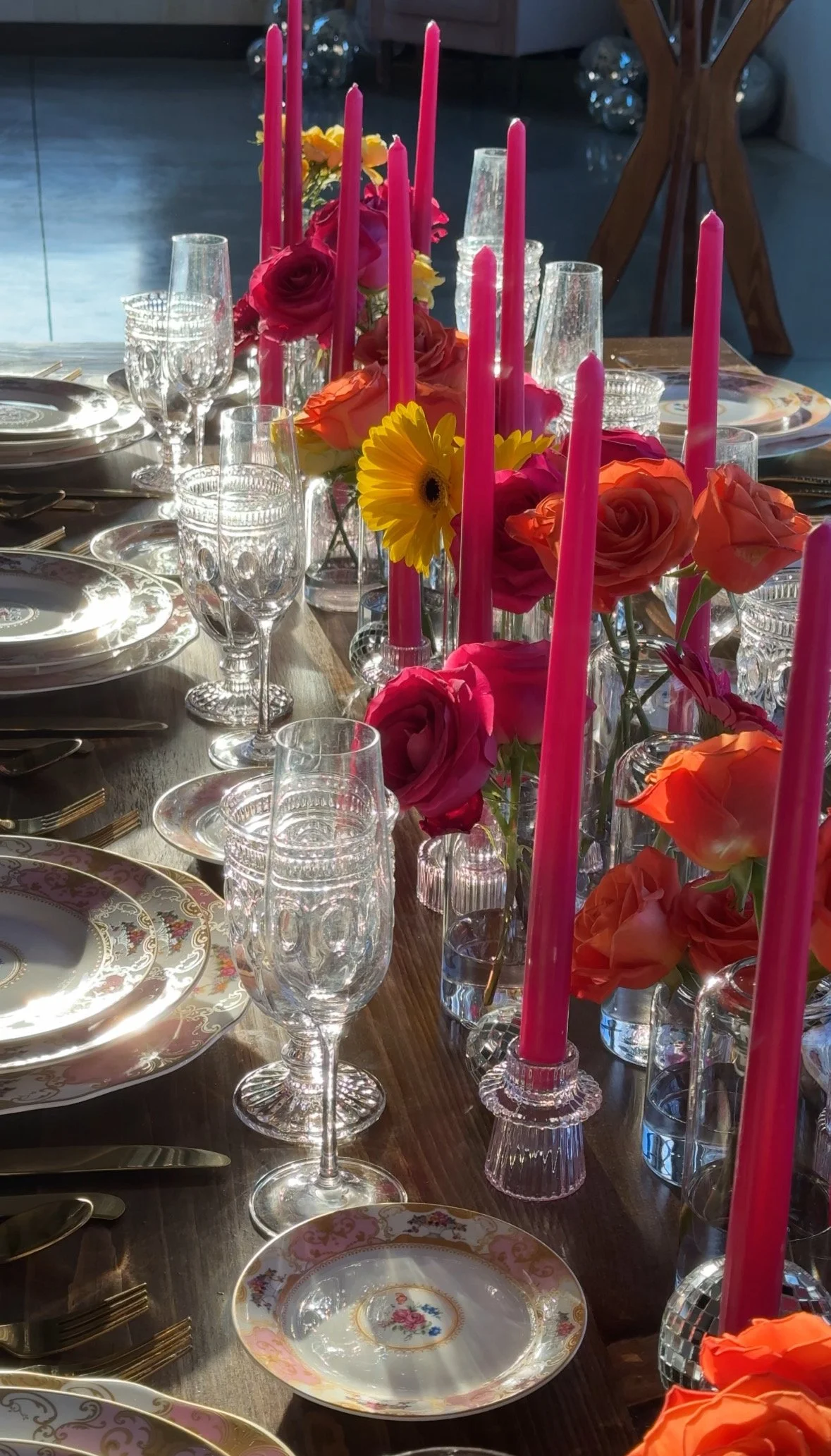 A decorated table set for a celebration with pink candles, colorful roses and flowers, clear glassware, and fine china plates.