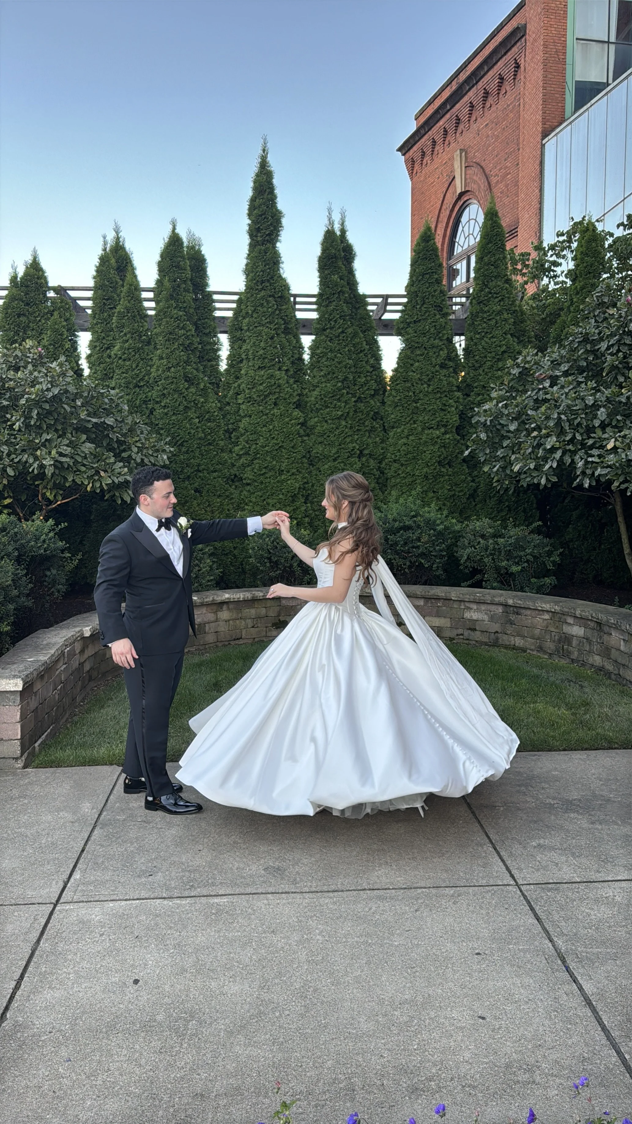 A bride and groom dancing outdoors in a garden area, with the bride wearing a white wedding gown and the groom in a black tuxedo. They are holding hands and looking at each other, with a backdrop of tall conifer trees, flowering bushes, and an old brick building.