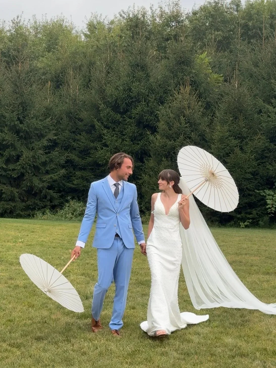 A newlywed couple holding umbrellas and walking hand in hand on a grassy field. The groom is wearing a light blue suit and the bride is in a white wedding dress with a long veil. They are surrounded by tall green trees.
