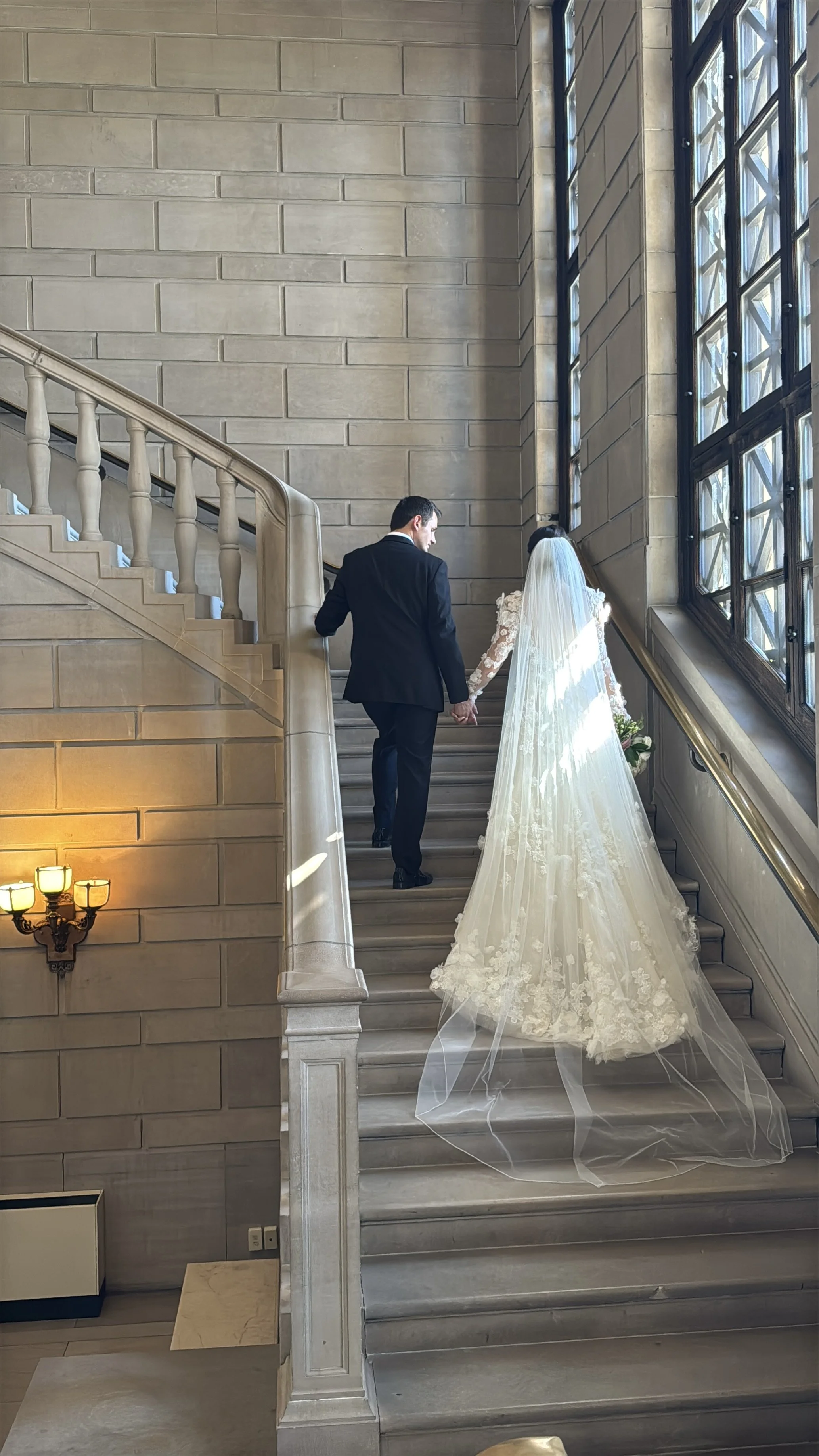 A newlywed couple walking up a stone staircase inside a classic building with large windows, the bride in a lace wedding dress with a long veil, holding hands and facing away from the camera.