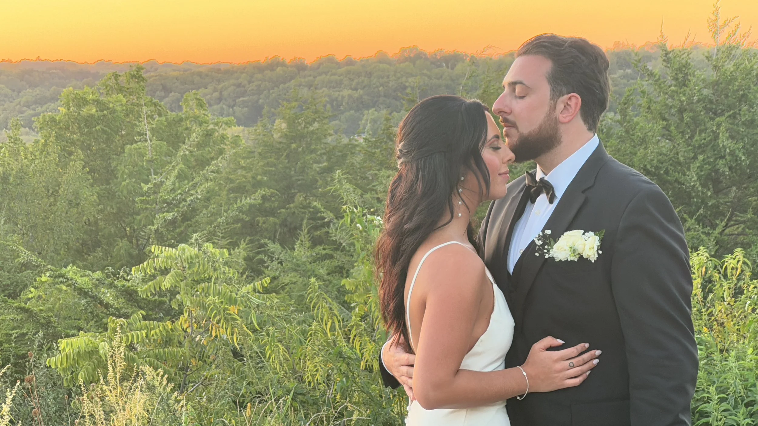 A couple dressed in wedding attire embracing in a lush, green outdoor setting at sunset, with a forest and colorful sky in the background.