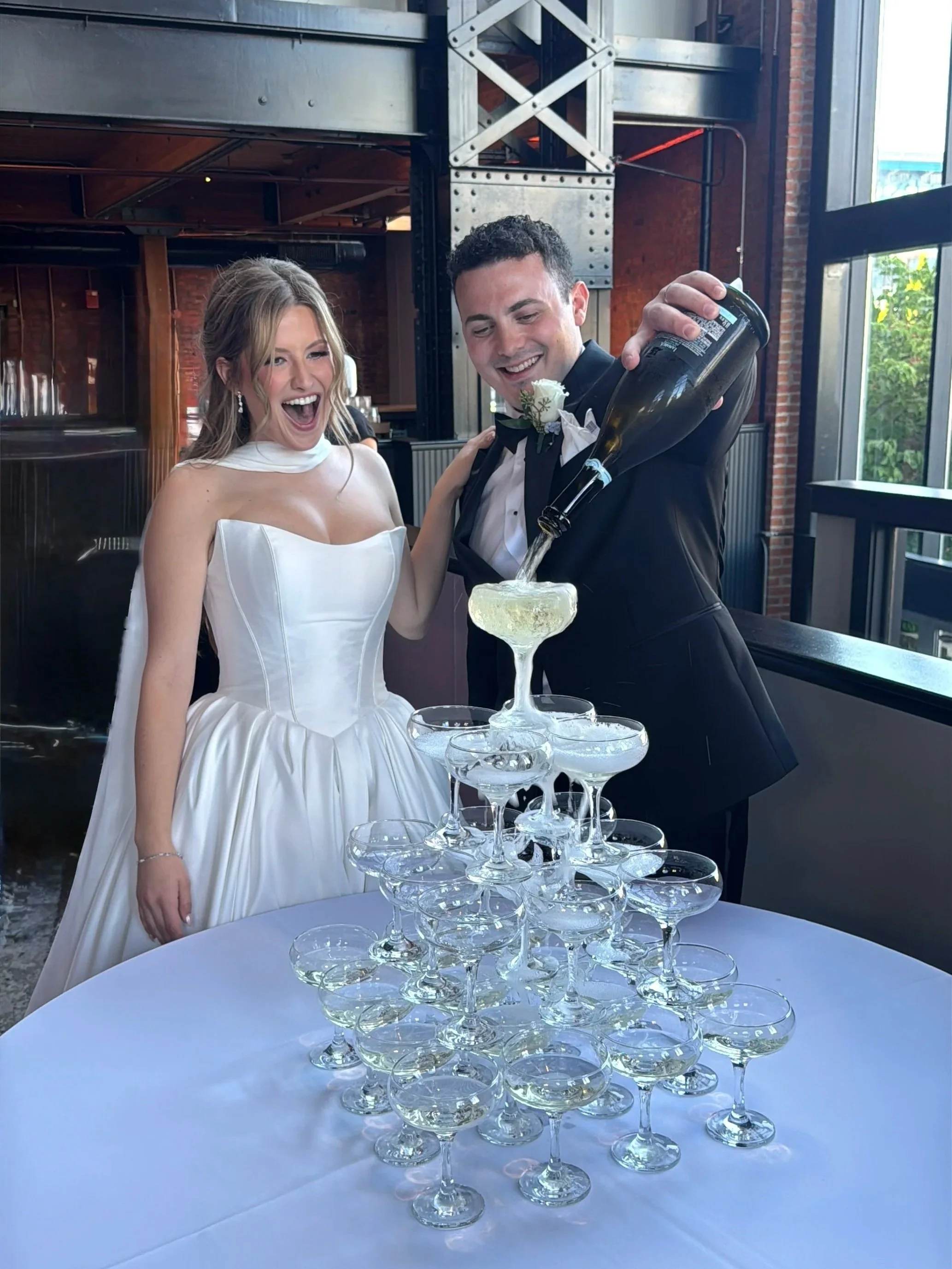 A bride and groom celebrating at a wedding reception, pouring champagne into a pyramid of glasses on a table, with a large window and industrial decor in the background.