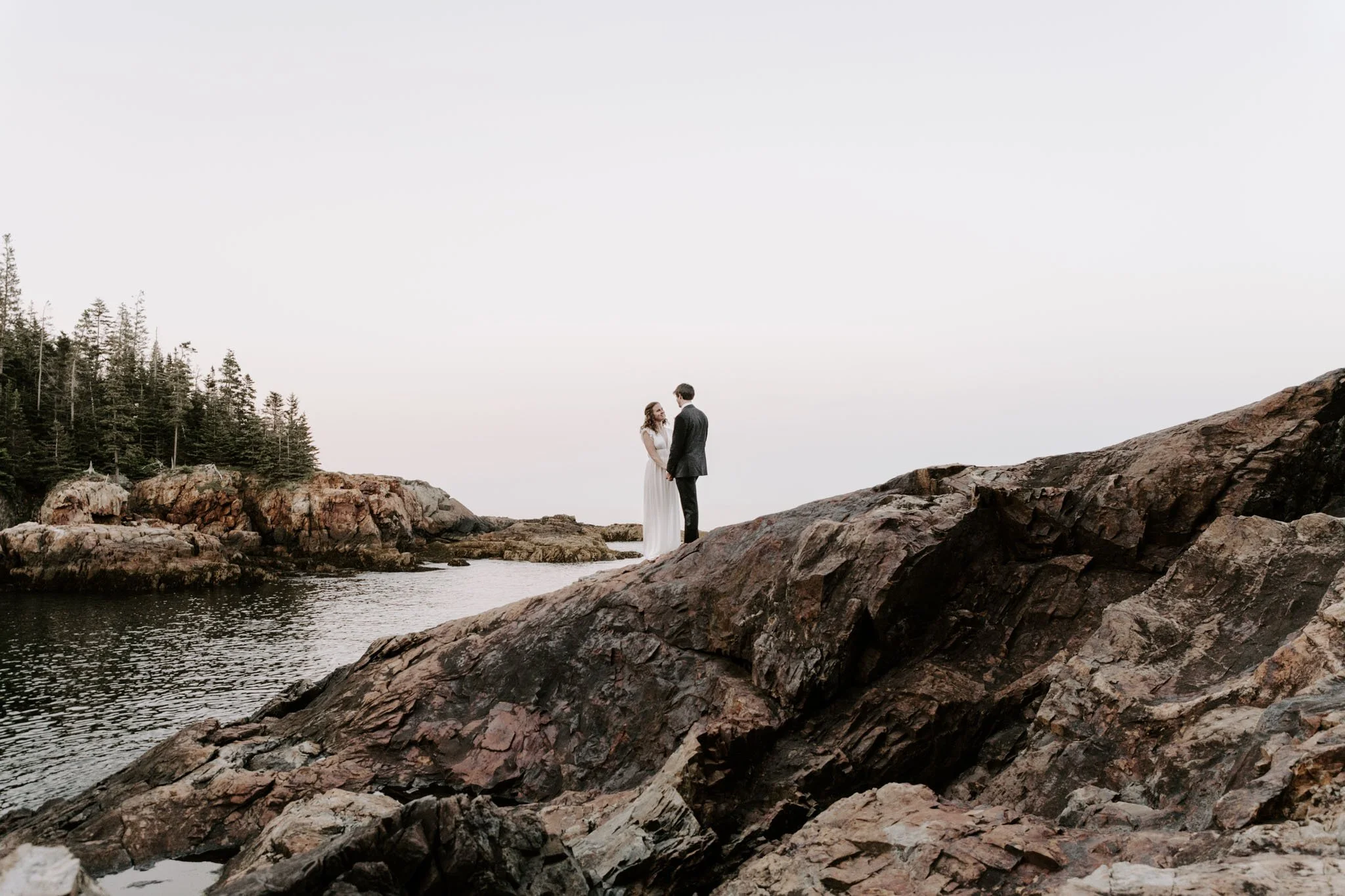 A couple in wedding attire holding hands on large rocks near water, with a forested shoreline in the background.