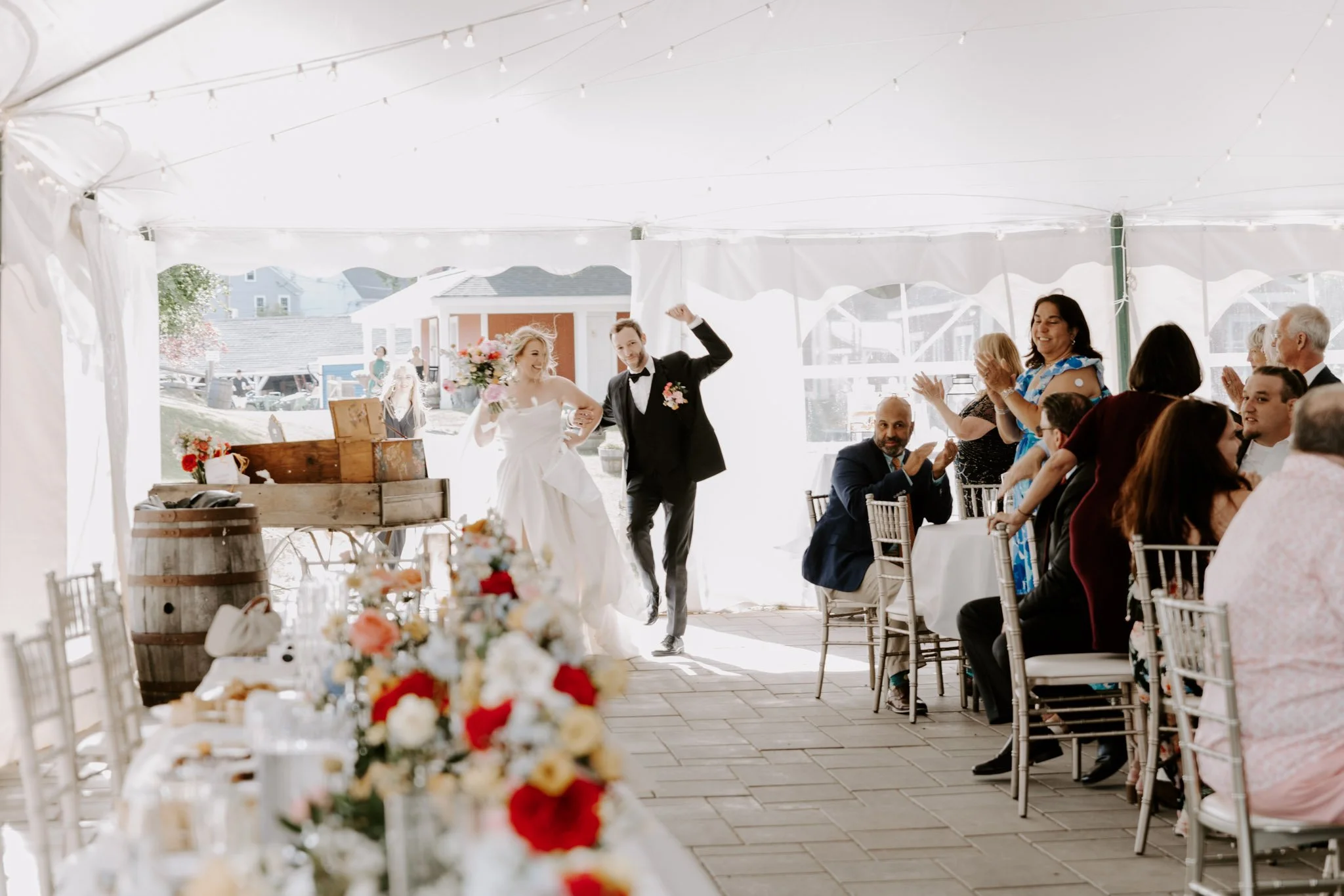 Bride and groom walking into a wedding reception tent with their guests applauding.