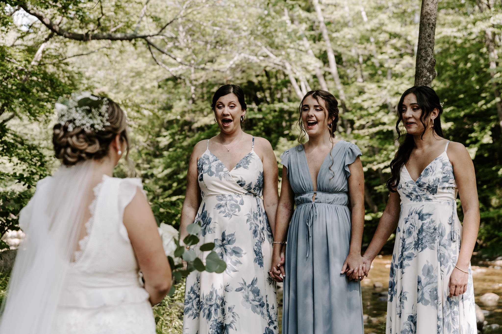A bride in a white wedding dress with lace details and a veil, holding a small bouquet, is facing three women holding hands, all dressed in blue and white floral dresses, outdoors in a green forest setting during a wedding ceremony.