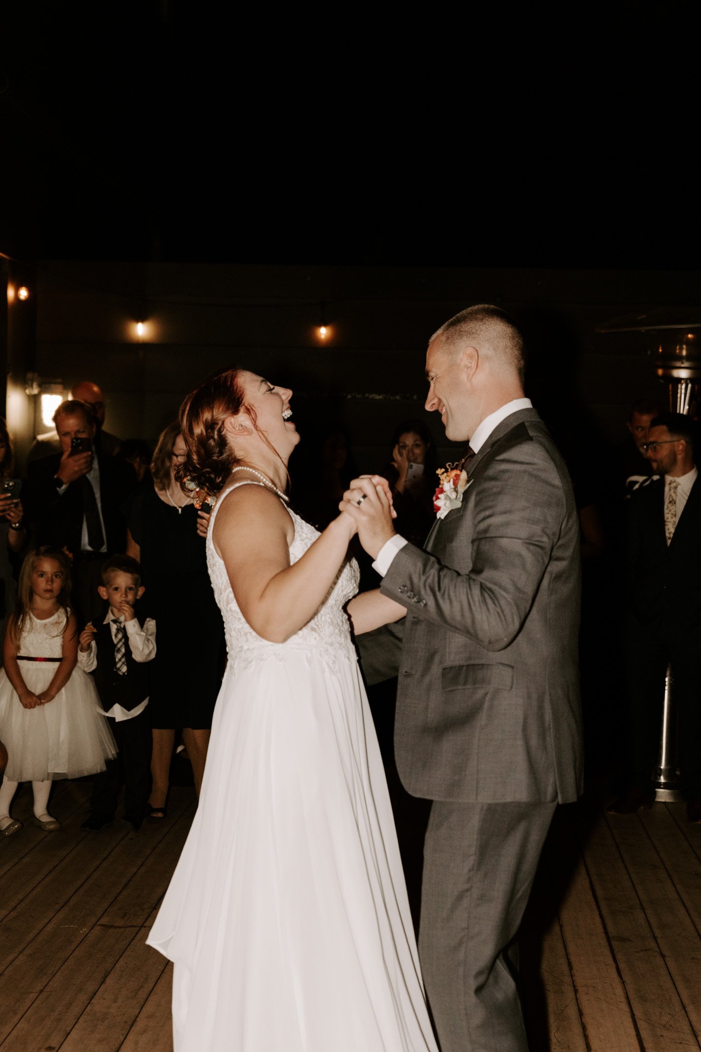 A bride and groom dancing at their wedding reception, smiling and holding hands, with guests watching and taking photos in the background.