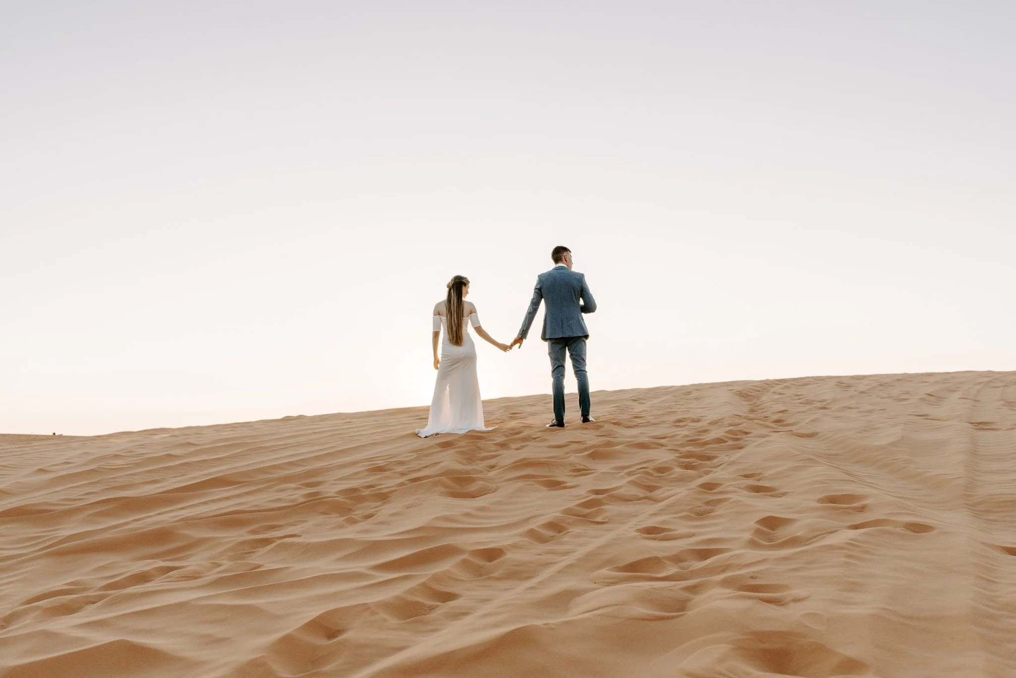 A man and woman in formal wedding attire holding hands and walking on sand dunes in a desert or beach area during sunset or sunrise.