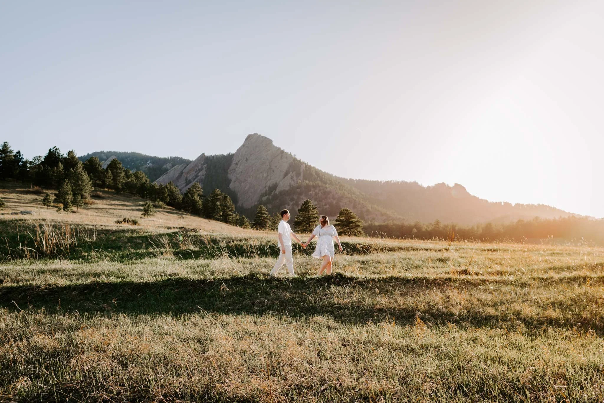 A couple holding hands and walking in a grassy field at sunset with mountains and trees in the background.