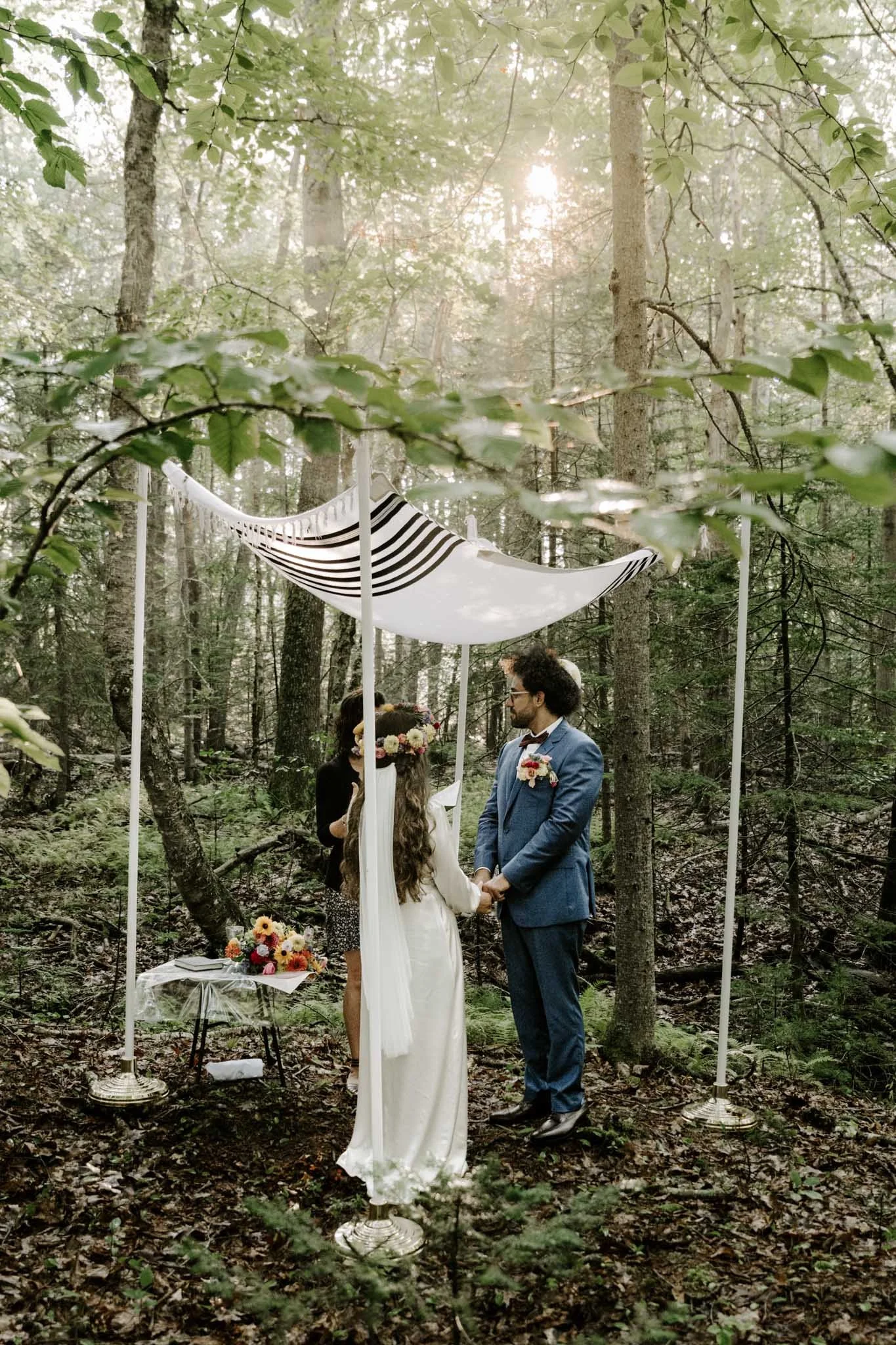 A couple getting married in a forest, holding hands under a striped canopy, with a woman officiating and a small table with flowers nearby.