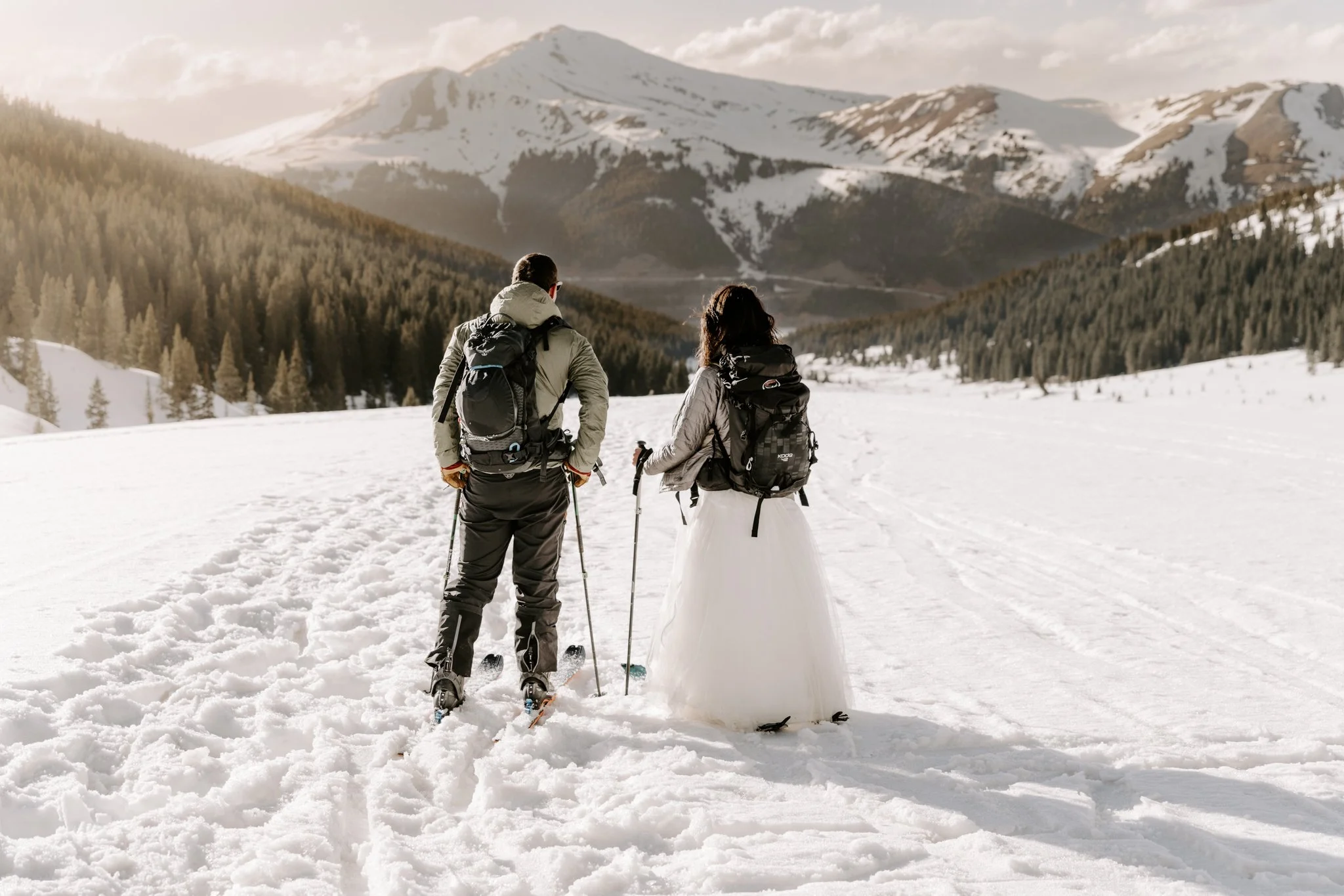 A couple dressed in winter clothes, wearing backpacks and ski gear, standing on snow in a mountain landscape with snow-capped peaks and pine trees in the background.