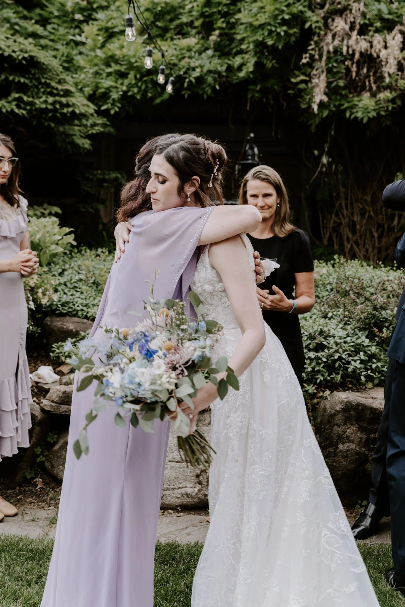 Two women hug at a wedding, one in a lavender dress holding a bouquet of flowers, embracing a bride in a white lace wedding gown, outside in a garden setting.