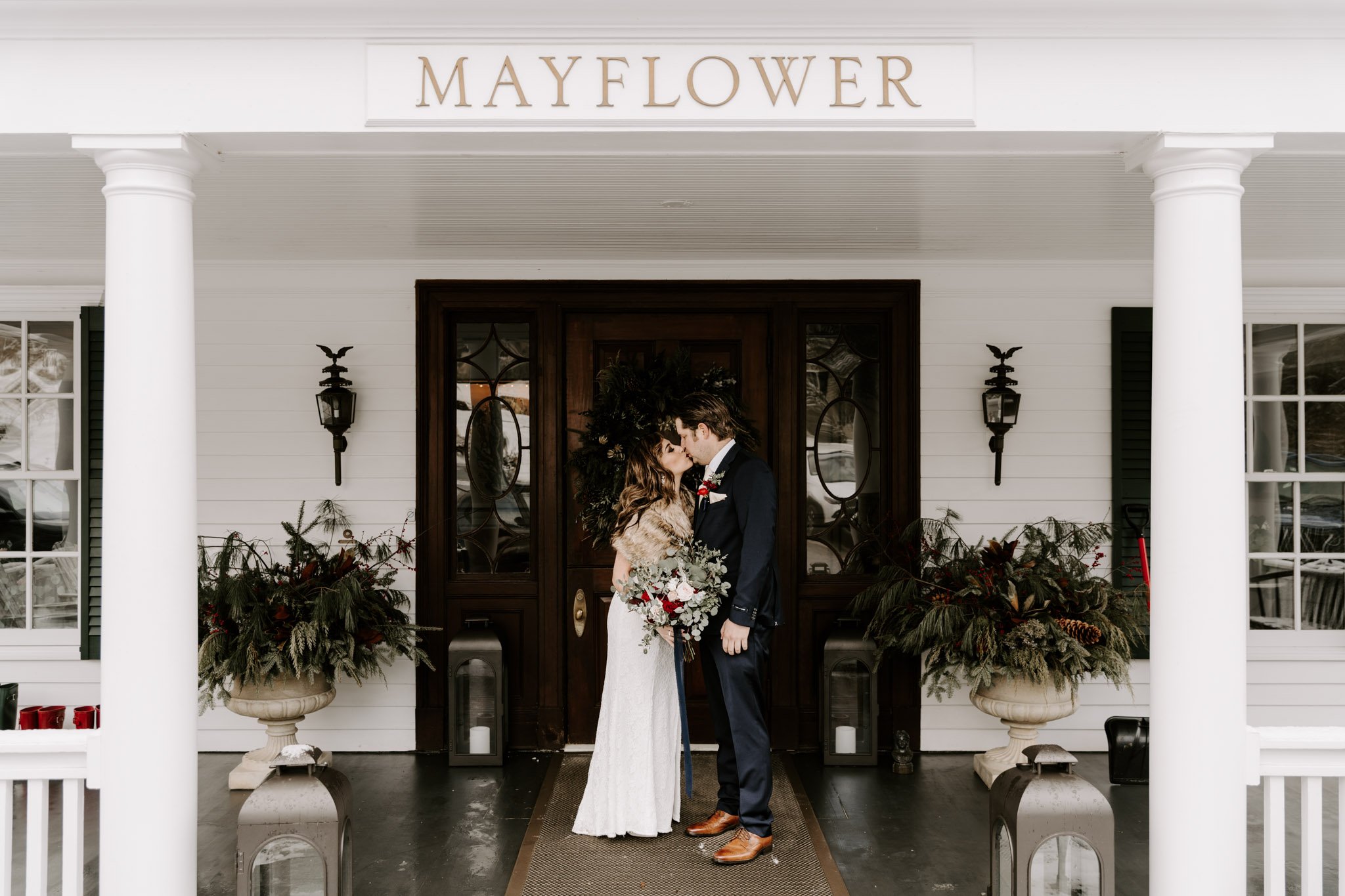 A bride and groom share a kiss in front of a white house with the sign read Mayflower. The bride wears a long white gown and fur shawl, holding a bouquet of flowers. The groom wears a dark suit, white shirt, and tie.