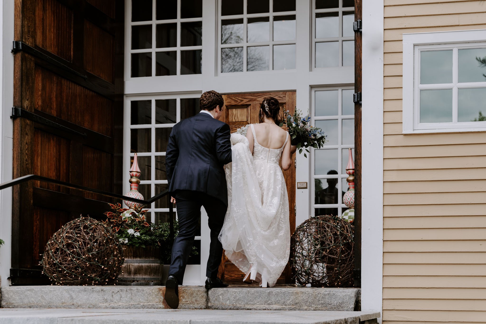 Bride and groom entering a house for a wedding, with the bride holding a bouquet and the groom in a blue suit.