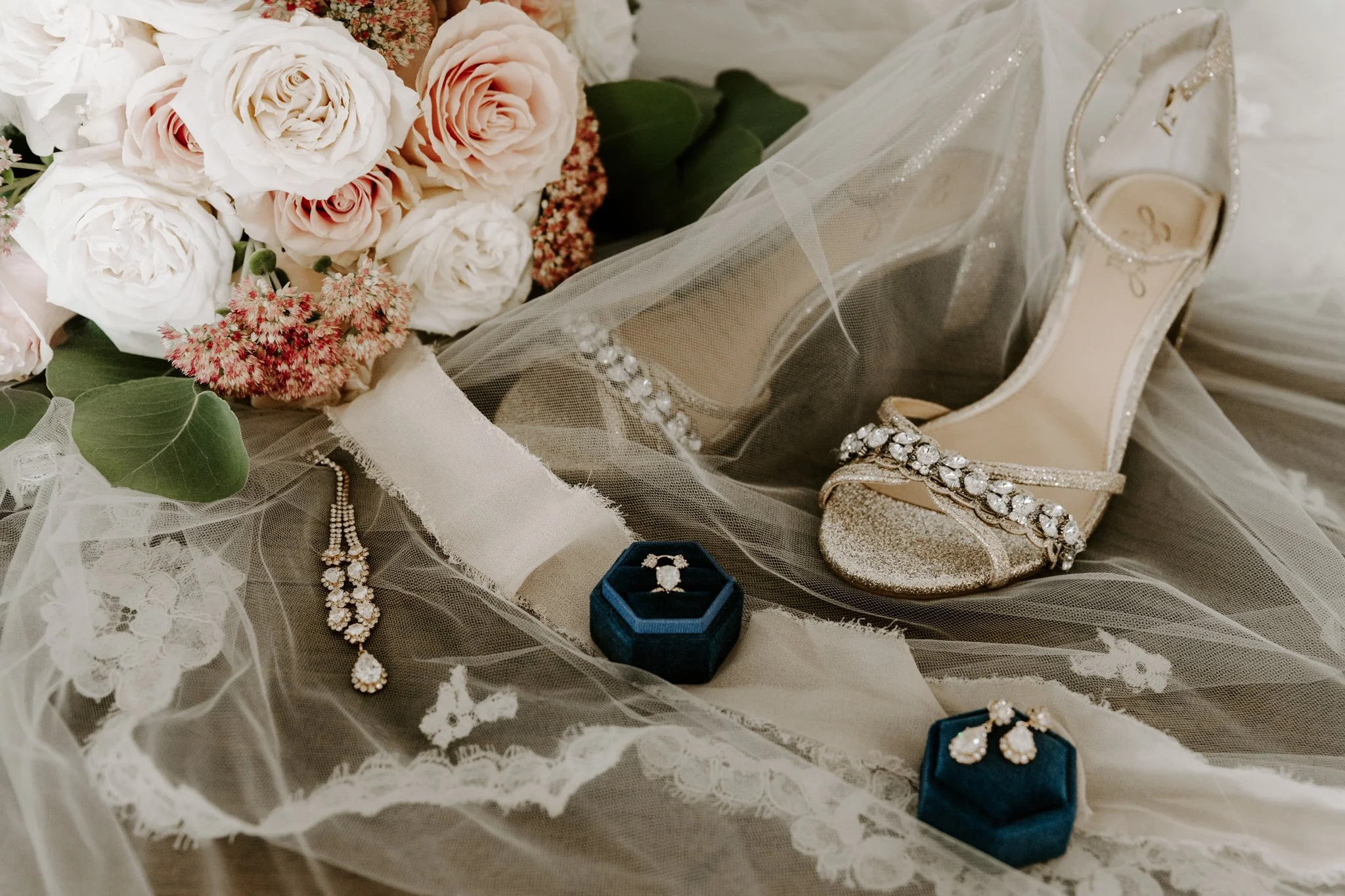 A collection of bridal accessories and flowers, including a bouquet of peach and white roses with green leaves, a pair of peep-toe high heels with rhinestone embellishments, a vintage engagement ring in a blue velvet box, matching earrings, and a pea