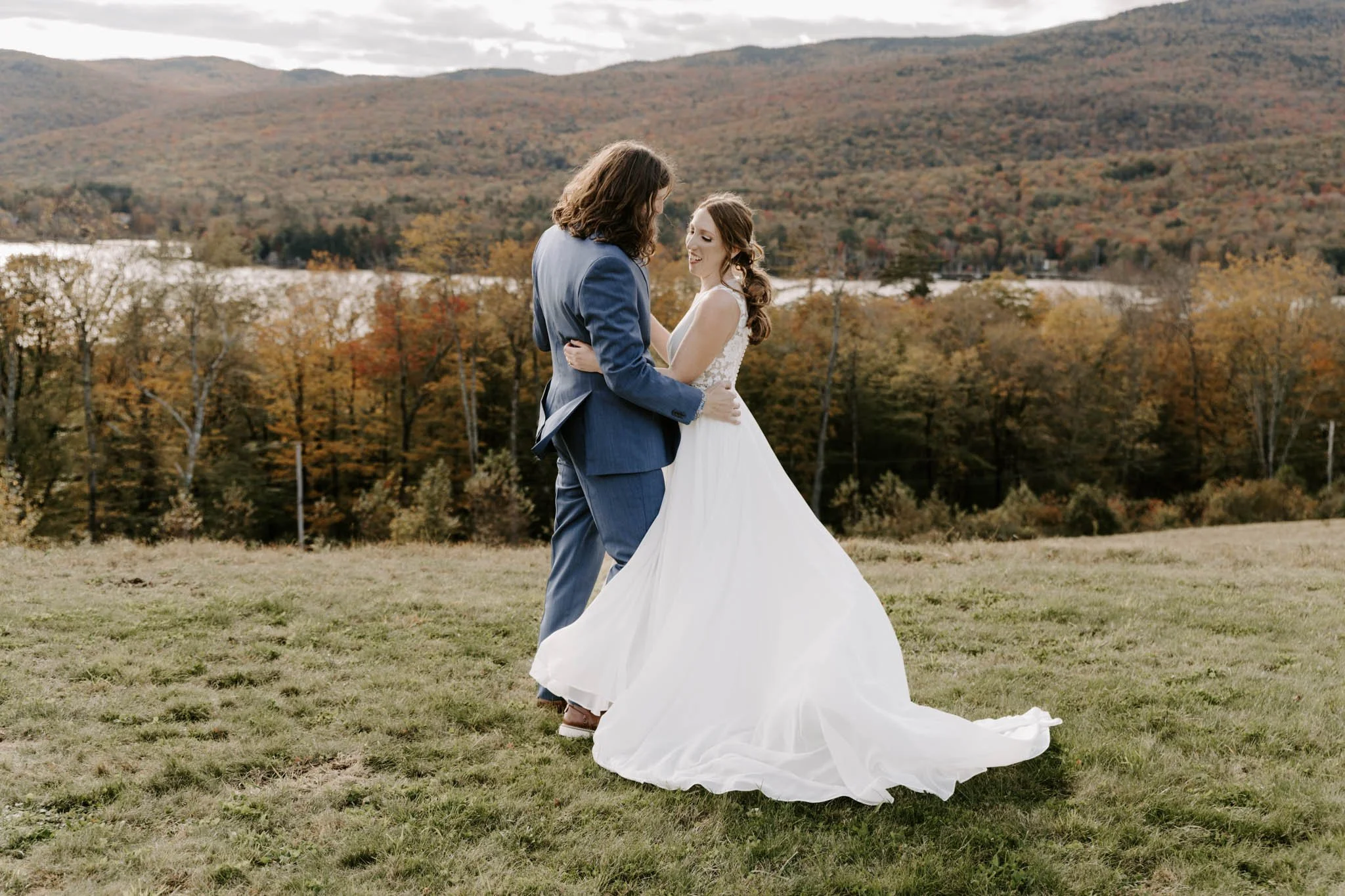 A bride and groom dance outdoors on a grassy field with mountains and trees with autumn foliage in the background.
