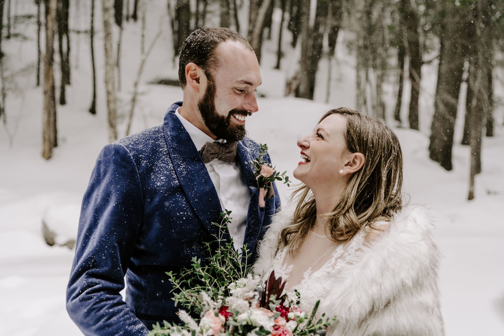 A couple in wedding attire sharing a joyful moment outdoors in a snowy forest, with the groom in a blue suit and the bride holding a bouquet of flowers.