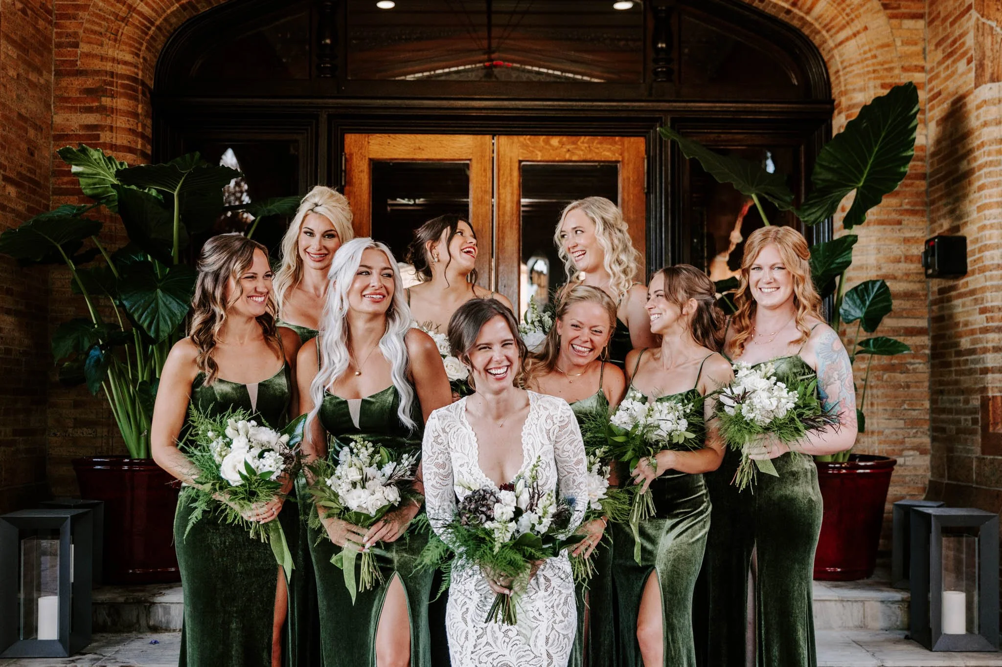 Bride with eight bridesmaids in green velvet dresses, holding bouquets, posing in front of wooden doors and brick walls, smiling and laughing.