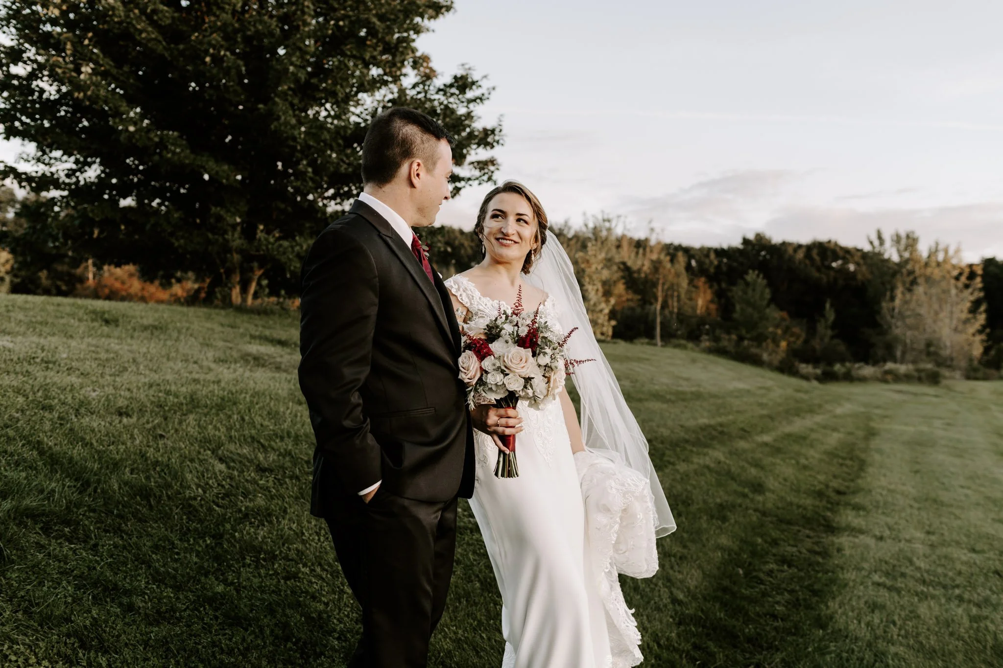 A bride and groom stand together outdoors on a grassy hill during sunset, smiling at each other. The bride wears a white wedding dress with a veil, holding a bouquet of roses and red foliage. The groom wears a black suit with a white shirt and red ti
