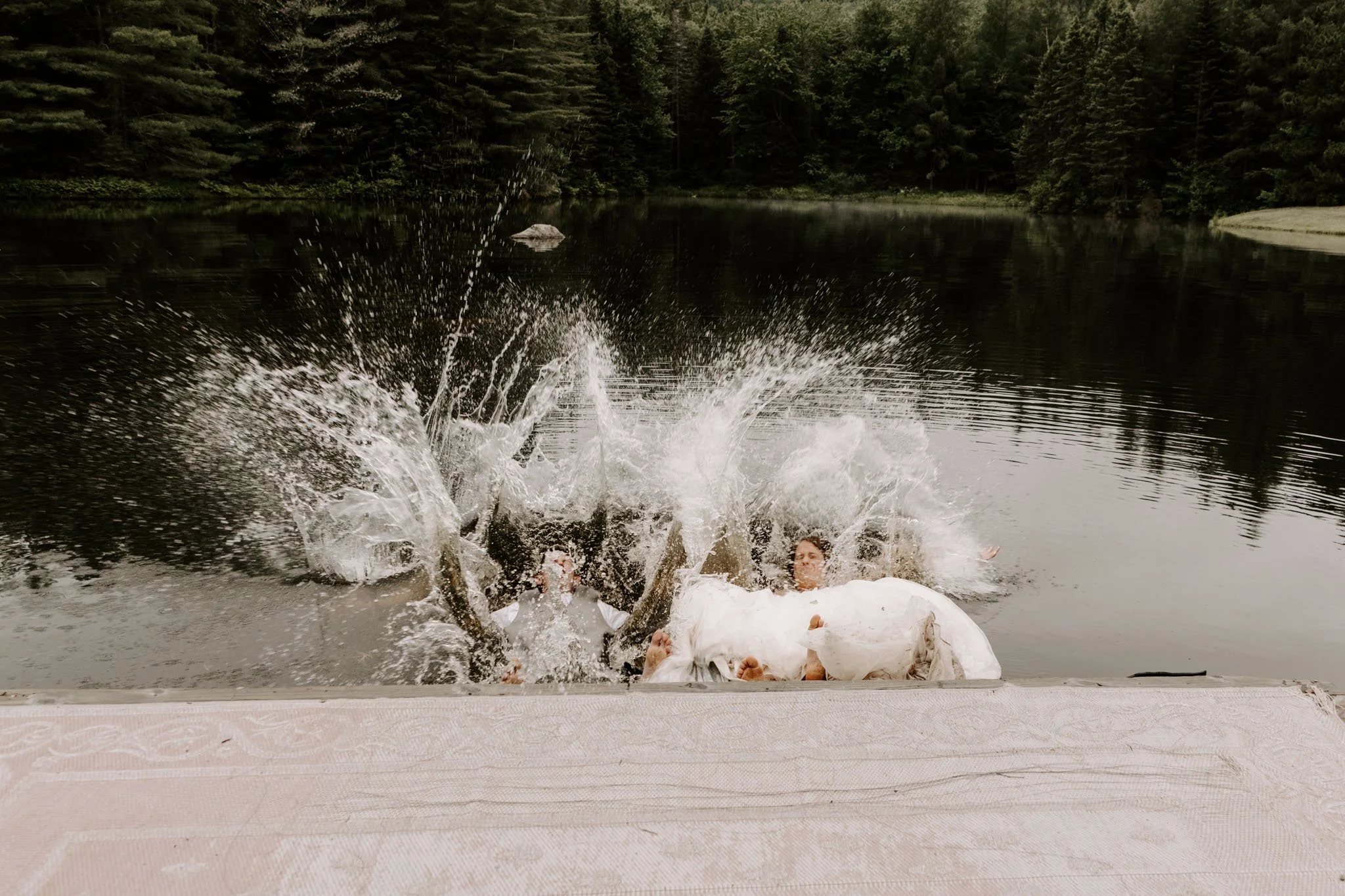 Two people in wedding attire falling into a lake, splashing water, from a dock with a forested background.
