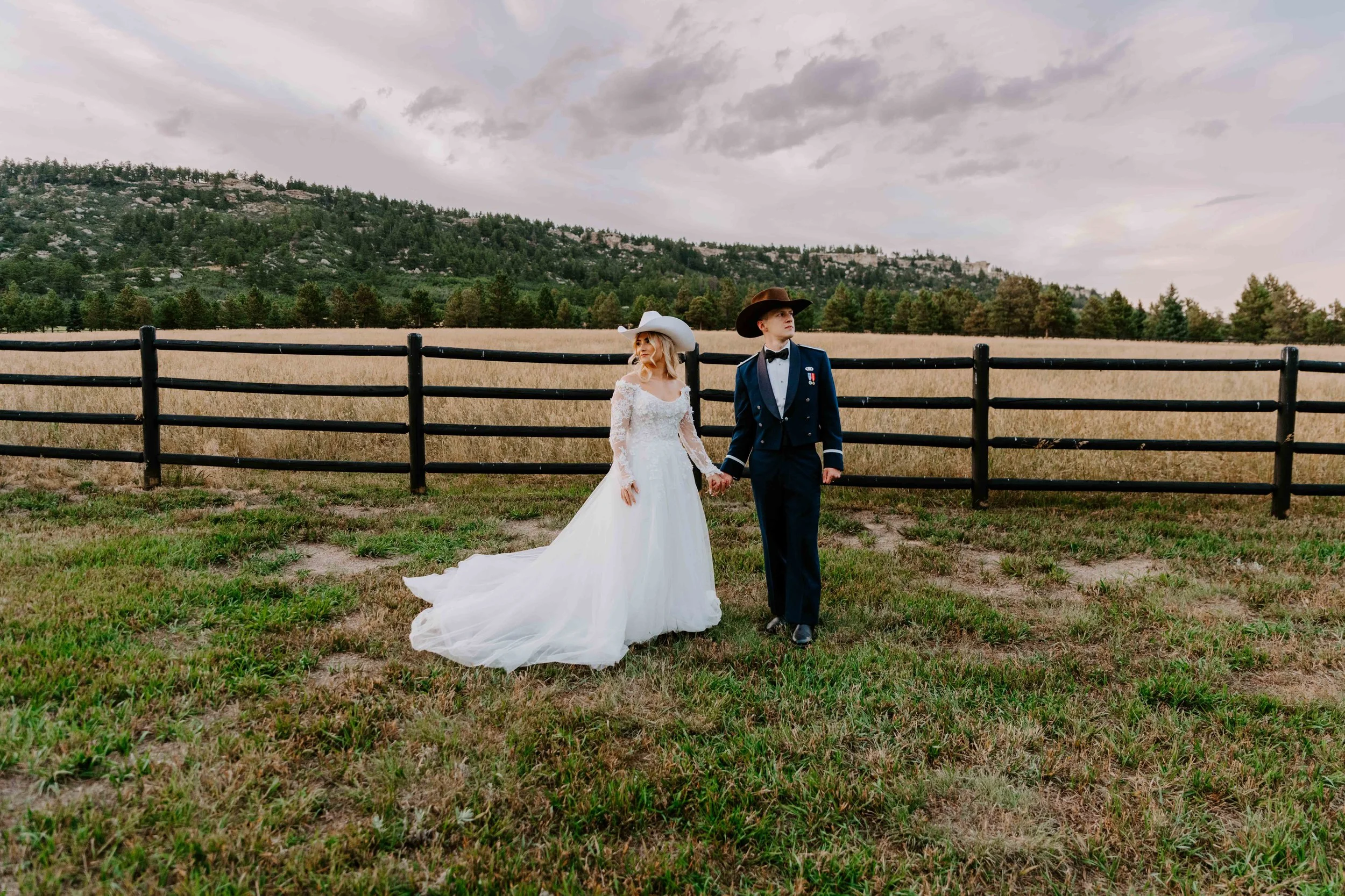 A couple dressed in wedding attire holding hands in an open field with a mountain backdrop and cloudy sky.