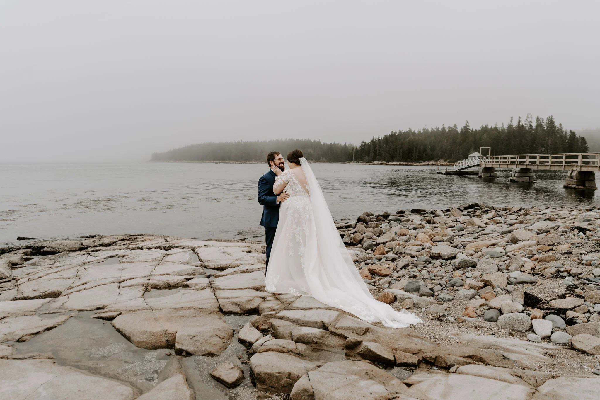 A bride and groom embrace on a rocky beach with foggy water and forested shoreline in the background.