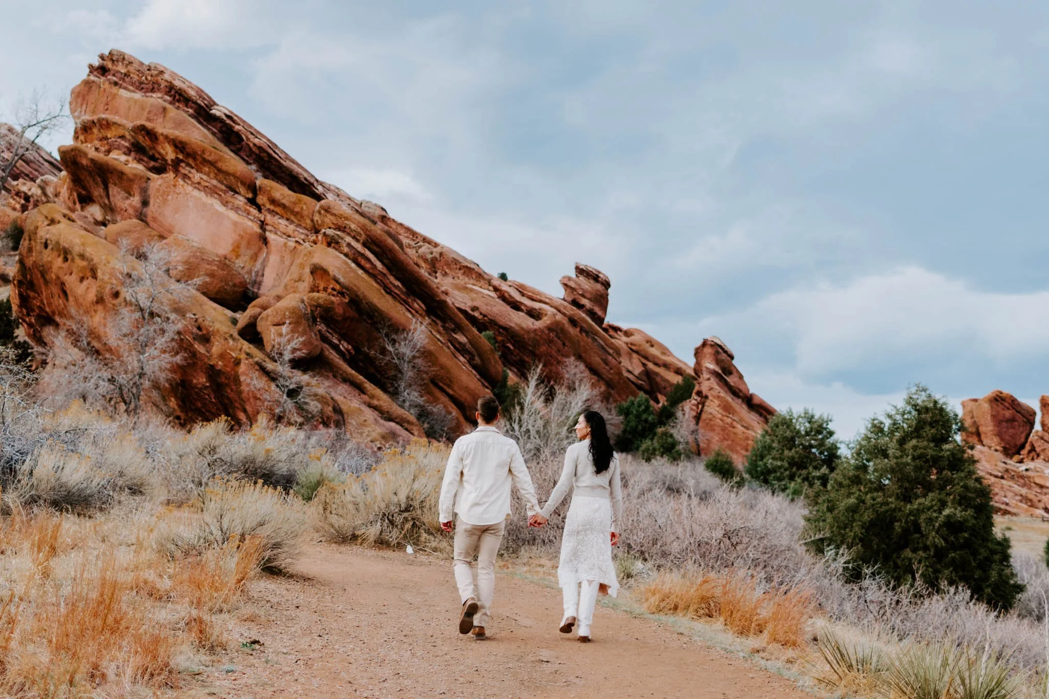 A couple dressed in white walking hand in hand on a dirt trail in a desert landscape with large red rock formations and sparse vegetation. Taken at Red Rocks in Colorado.