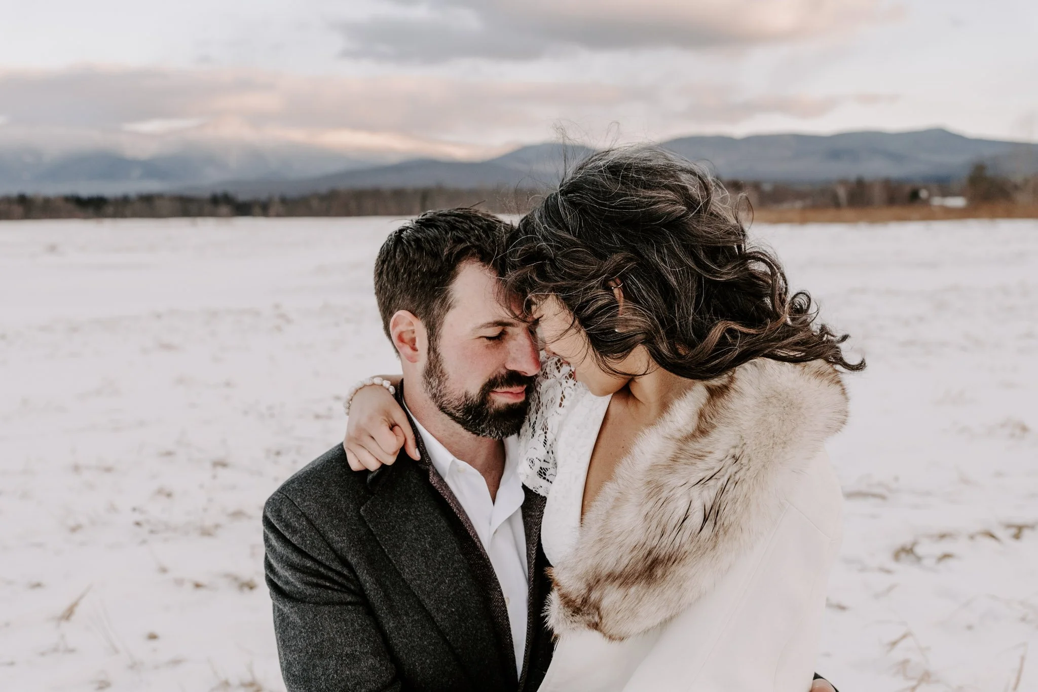 A couple standing close together in a snowy field with mountains in the background, faces touching.