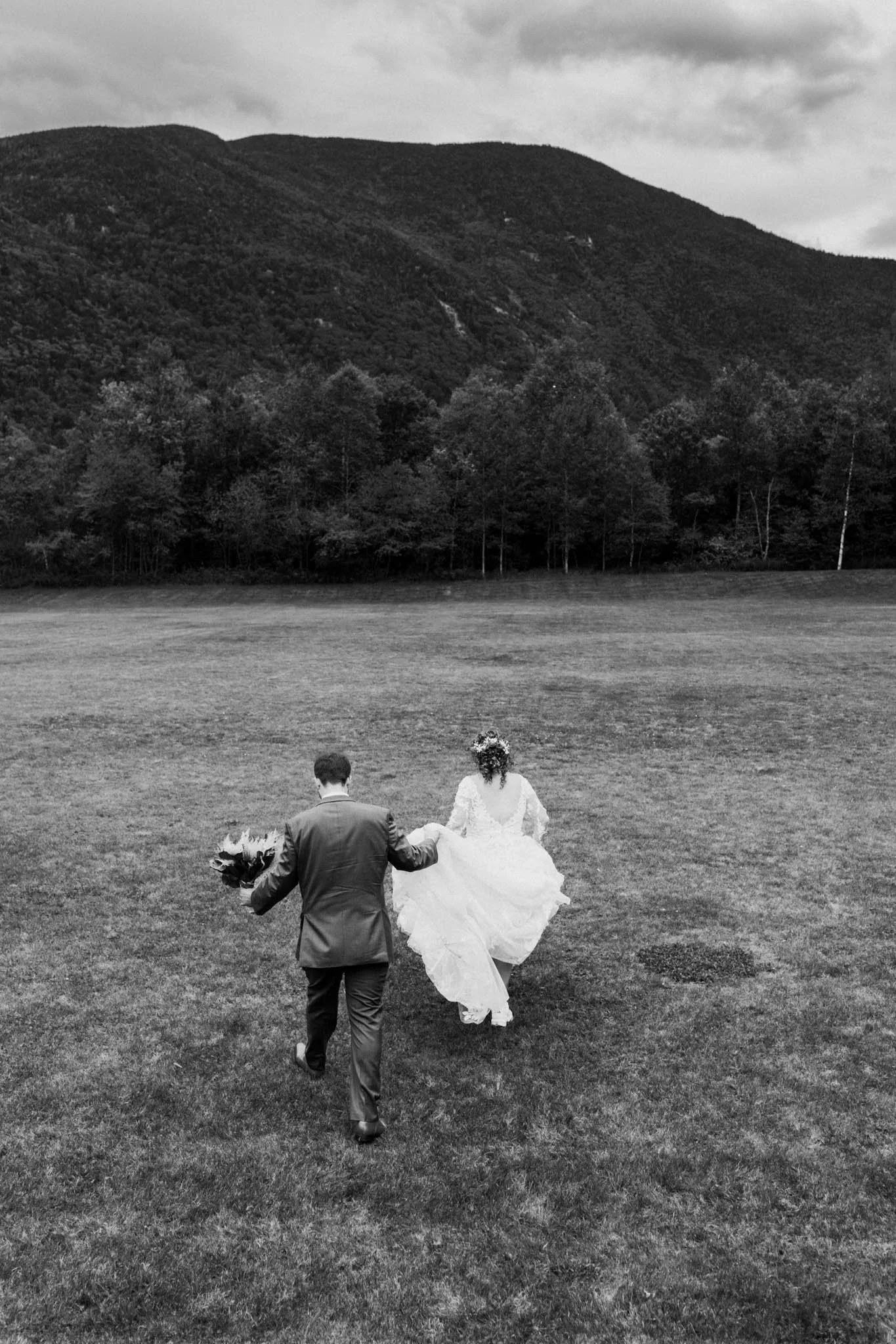 A black and white photo of a wedding couple walking across a large open grassy field with mountains and trees in the background. The bride is holding her dress as she walks, and the groom is holding a bouquet of flowers.