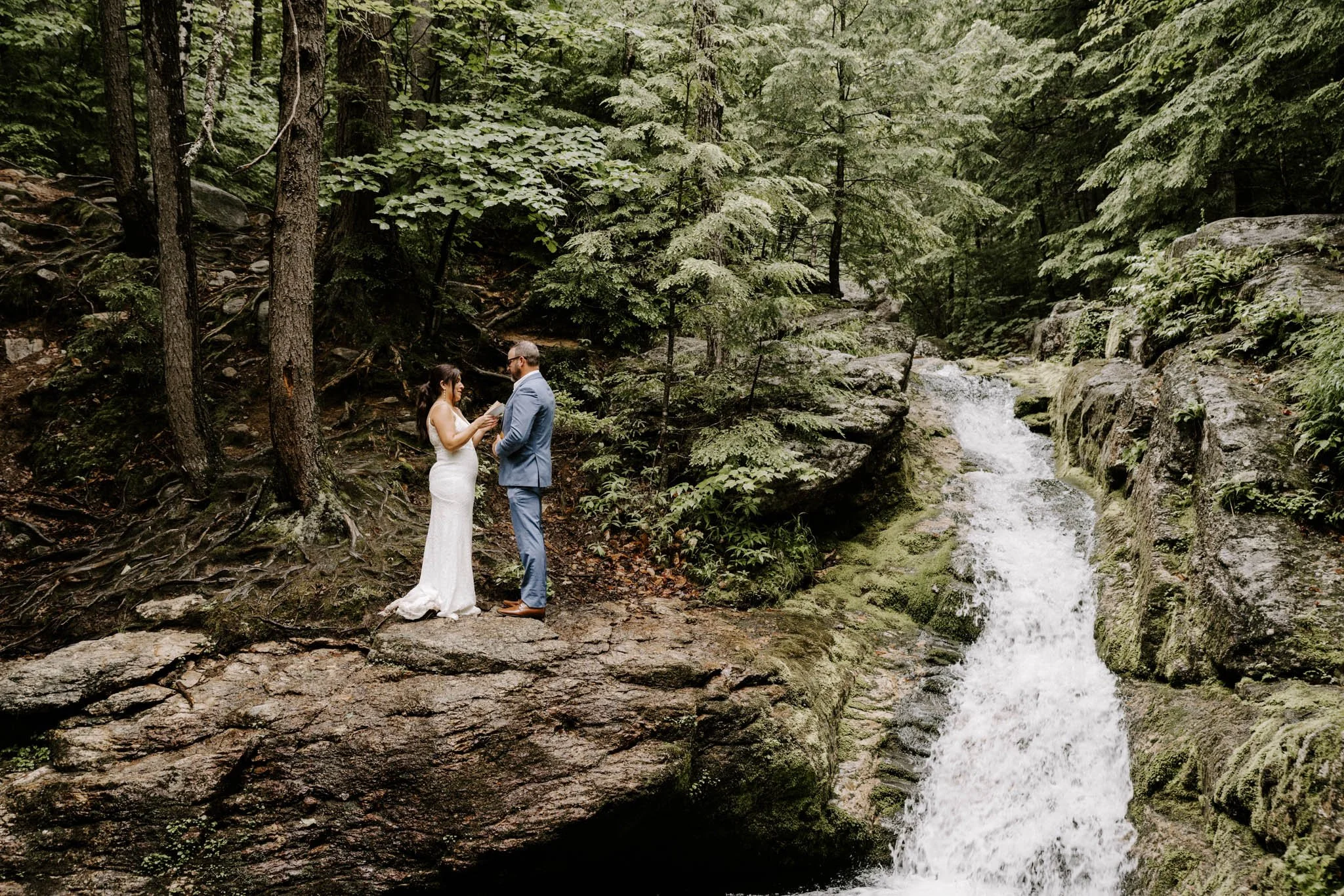 A couple dressed in wedding attire standing on a large rock in front of a waterfall in a forest.