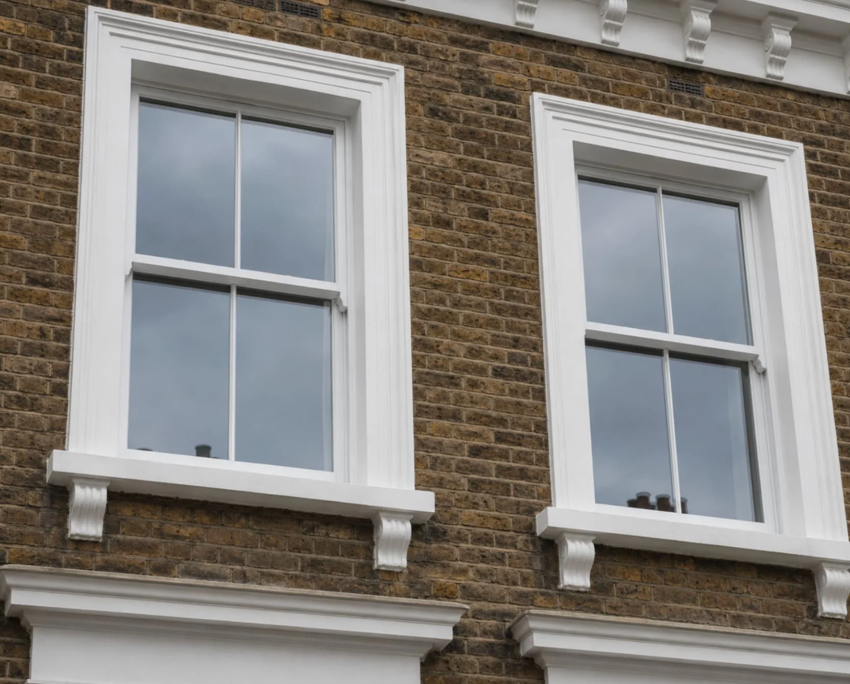 Two white-framed, double-hung sash windows on a brick building, with decorative white trim and molding, reflecting cloudy sky.