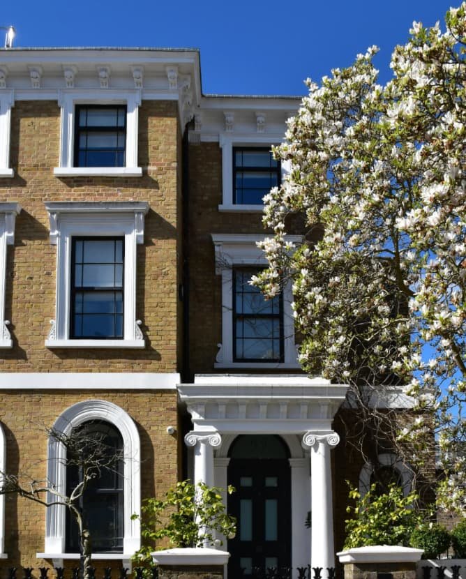 Victorian brick townhouse with white decorative trims, black window frames, and a black front door, surrounded by blooming trees under a clear blue sky.