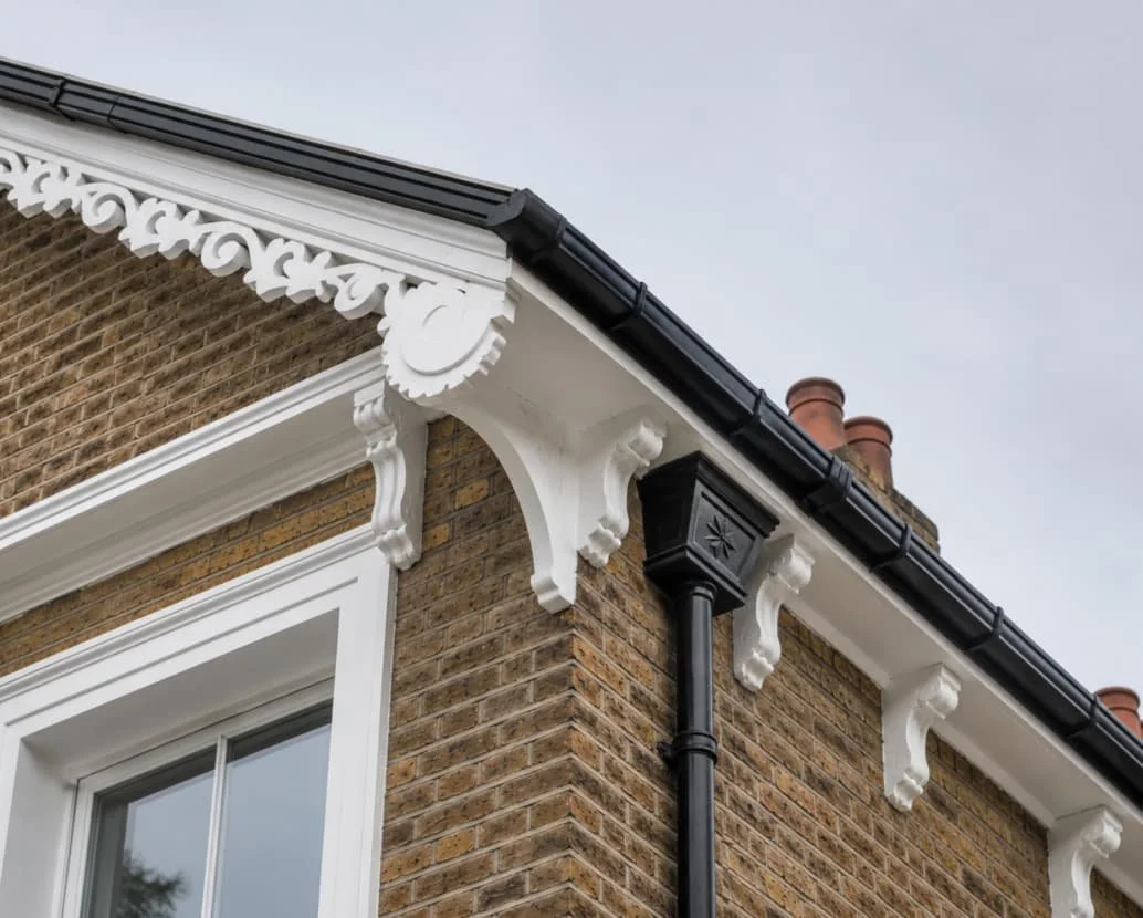 Close-up of the corner of a brick house showing ornate white decorative woodwork under the black roof gutter, a black drainpipe with a decorative star pattern, and two brick chimneys in the background against an overcast sky.