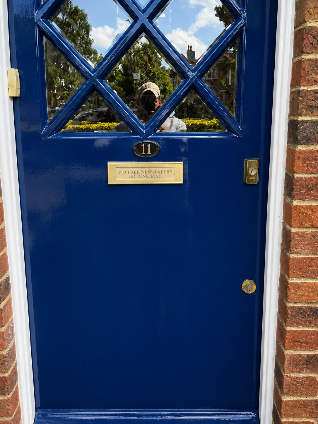 Close-up of a blue front door with a windowed upper section, brick wall on the right, and a brass mailbox plate with the number 11 and a sign that says 'No free newspapers or junk mail.' Reflection of a person taking the photo is visible in the window.