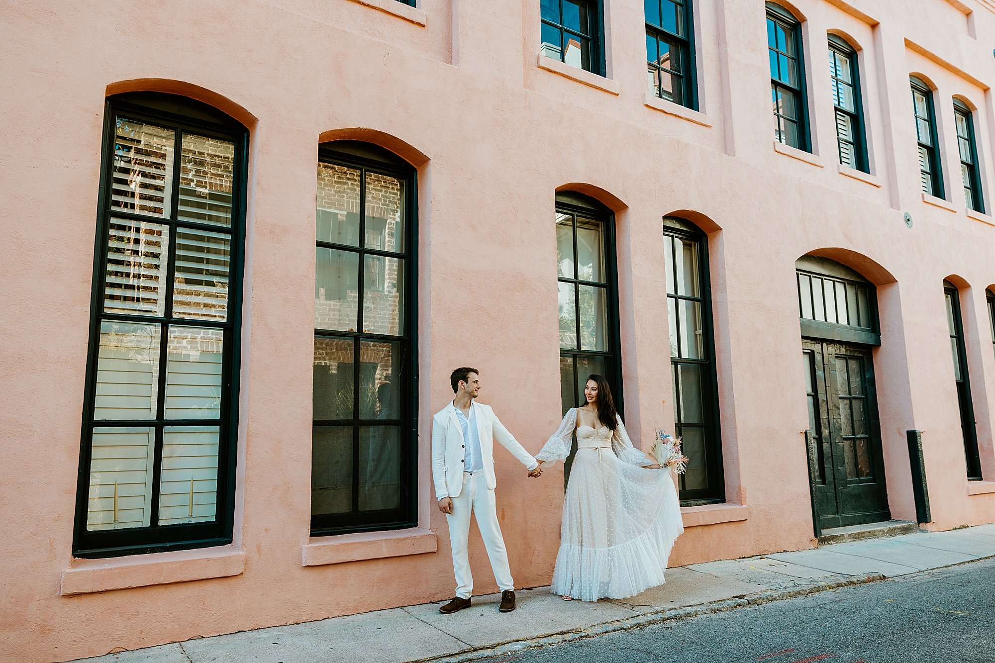 How to elope in Charleston SC - couple walks in front of a very large pink house with old historic windows