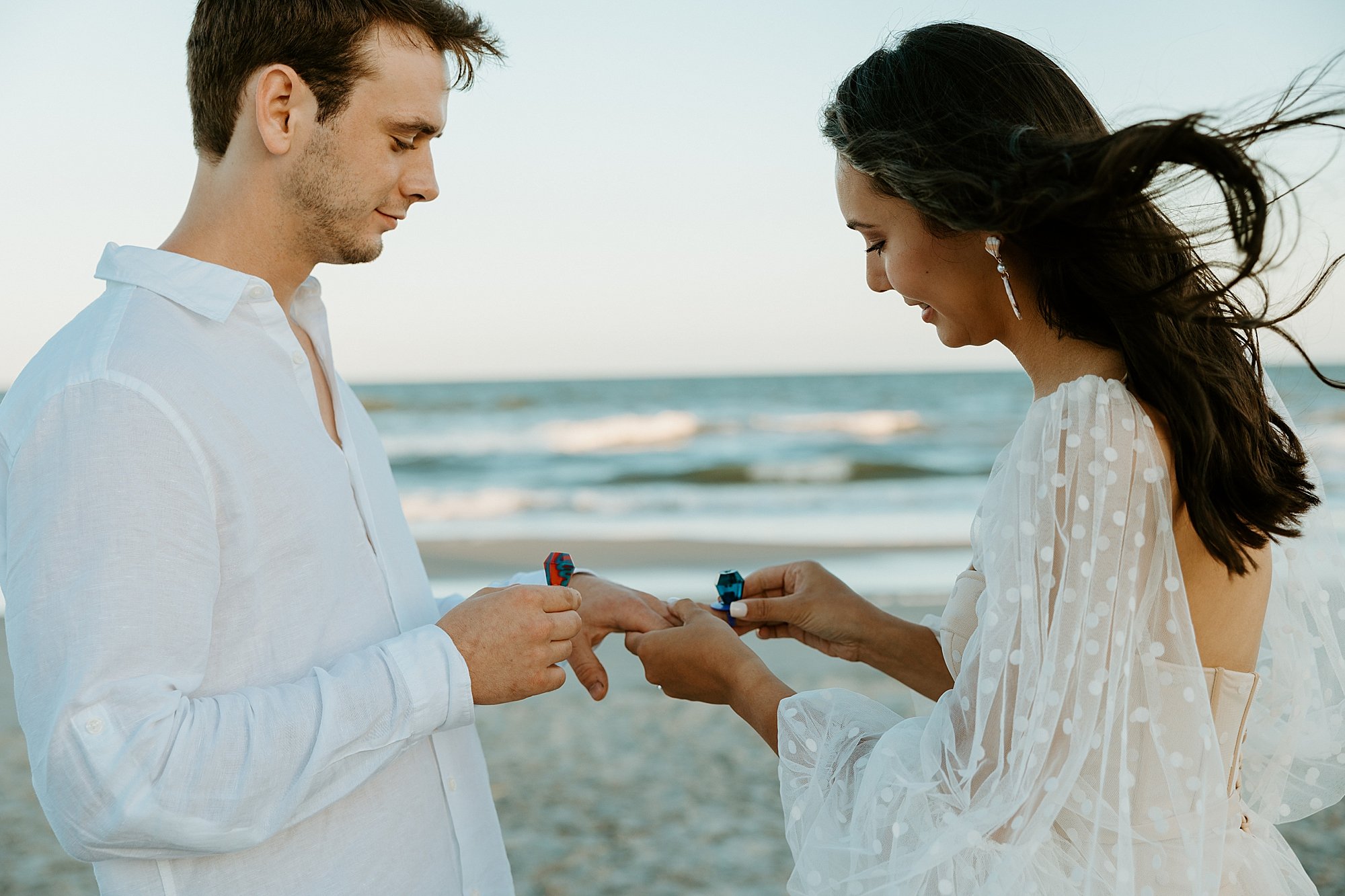 Couple exchanges ring pops on the beach as a symbol of their marriage. The sky is starting to turn pink because of the sunset.