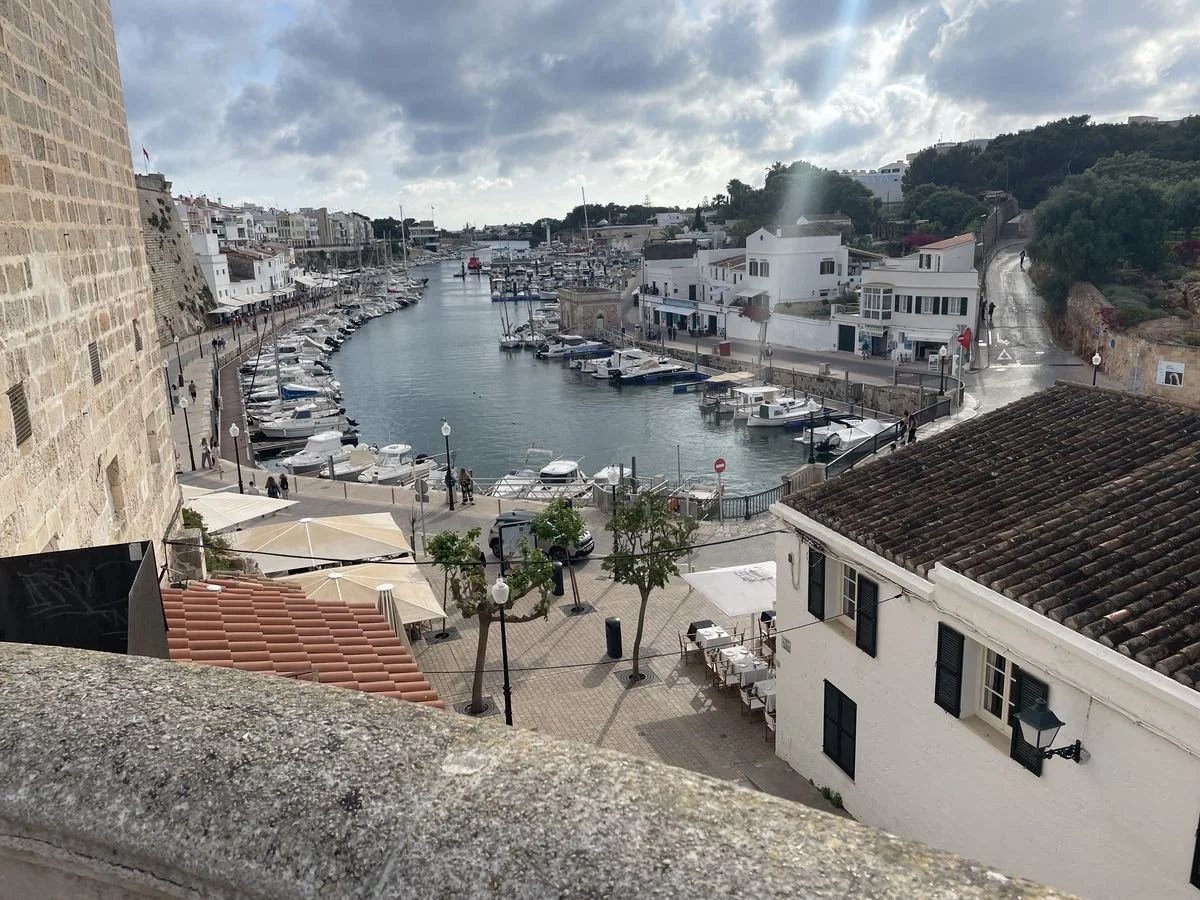 A picturesque coastal scene of a small marina with boats docked, surrounded by white buildings and narrow streets, under a cloudy sky.