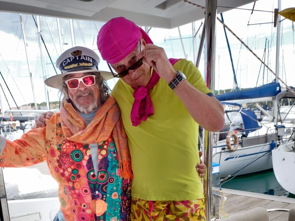 Two men on a boat posing for a photo, one wearing a captain's hat and colorful shirt, the other in a pink bandana and yellow t-shirt, with sailboats in the background.