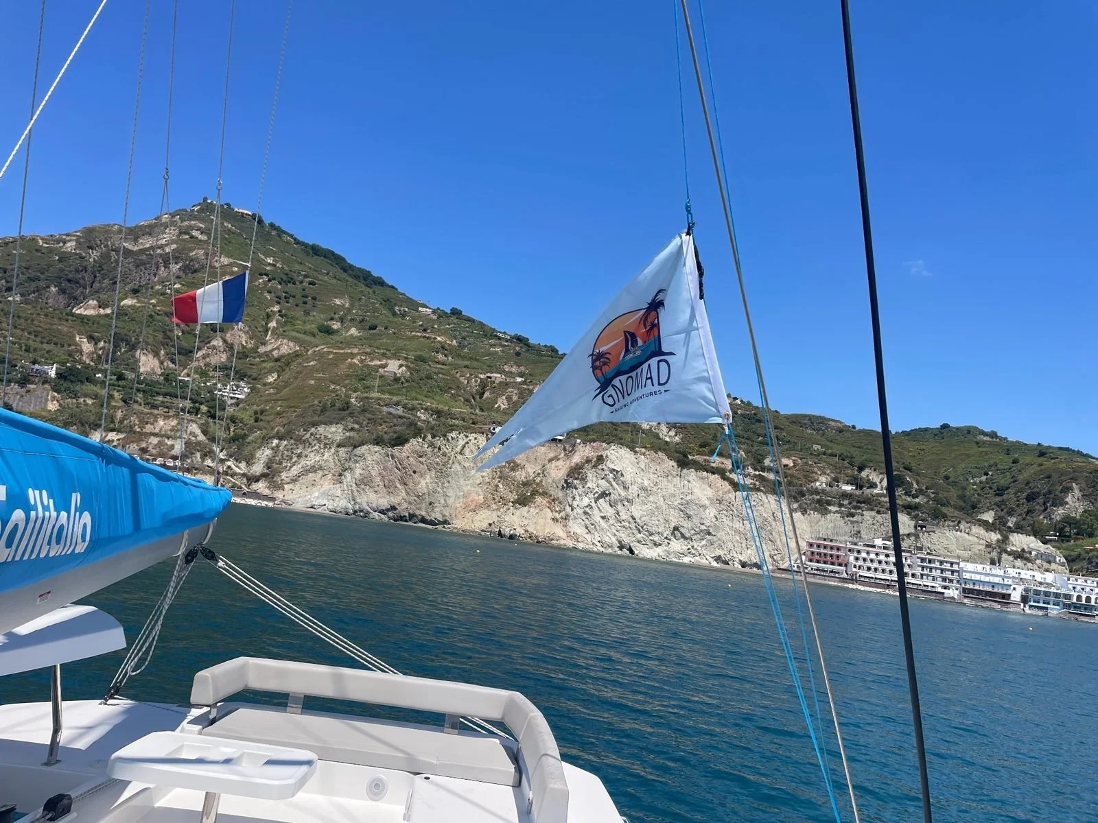 View from a sailboat with a white sail and a flag that reads 'Gnomad Sailing Adventures,' on a body of water near a rocky coastline with hillside houses and a clear blue sky.